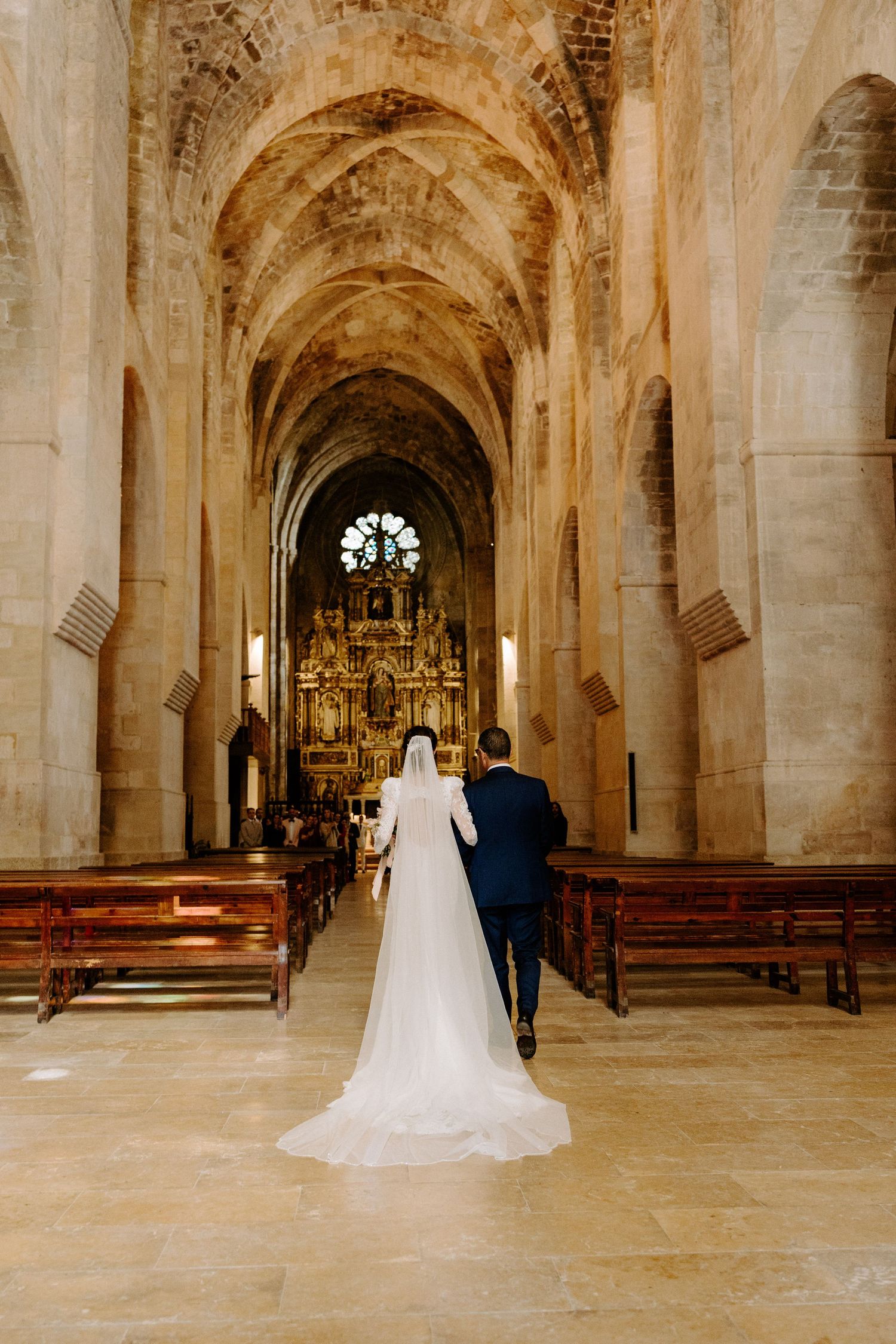 A couple walks down the aisle of a grand cathedral with vaulted ceilings and ornate golden altar during their wedding ceremony.