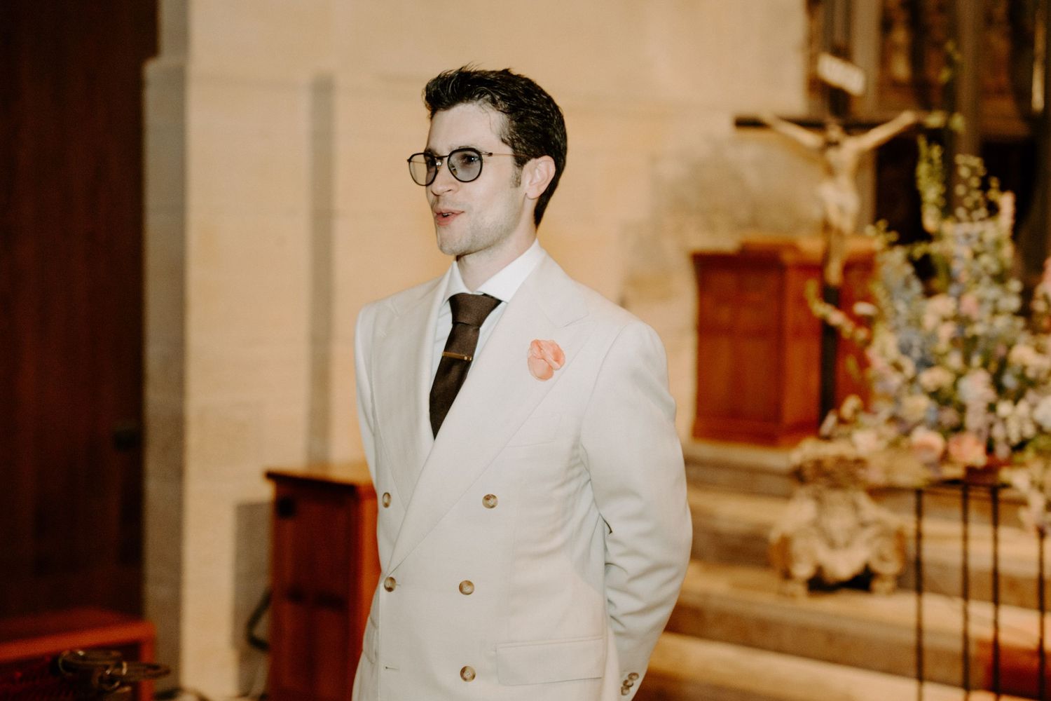A person in a white double-breasted suit and glasses stands in an elegant church setting.