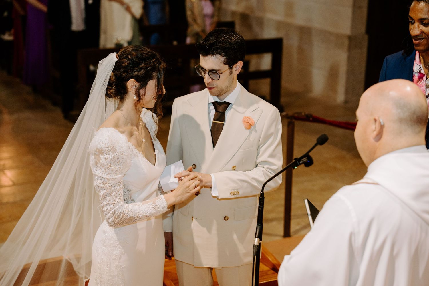 Couple exchanges wedding rings during ceremony as priest presides in historic church interior.