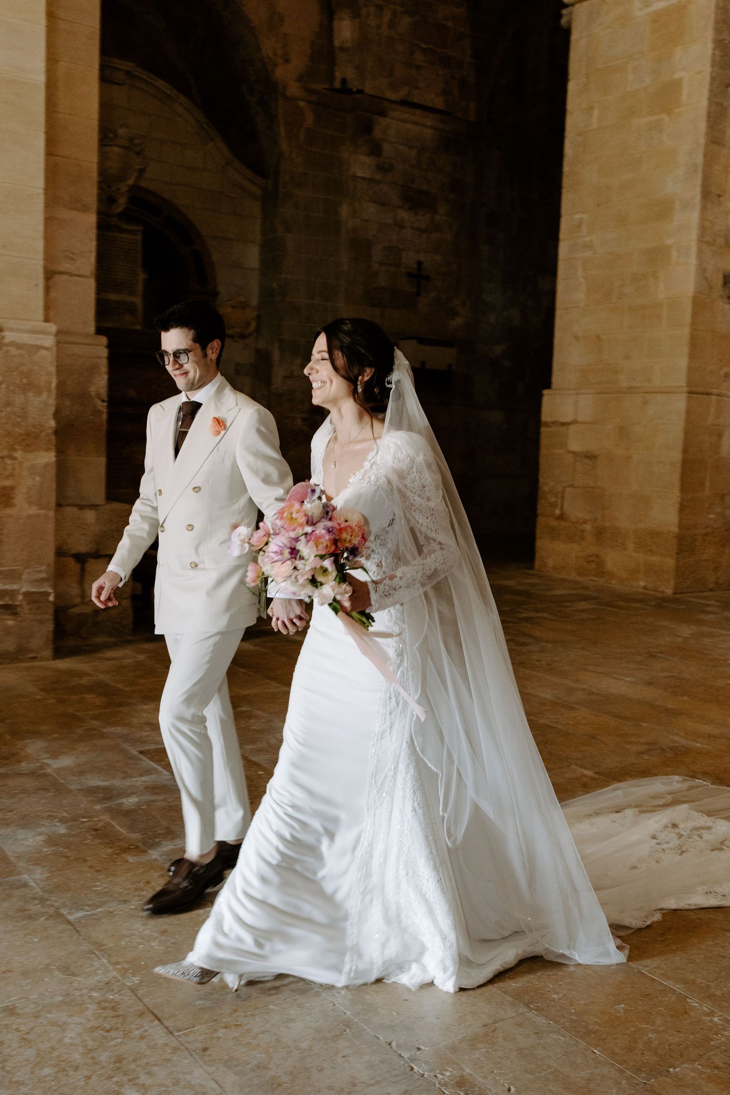 Wedding couple walks hand in hand through a grand stone church interior.