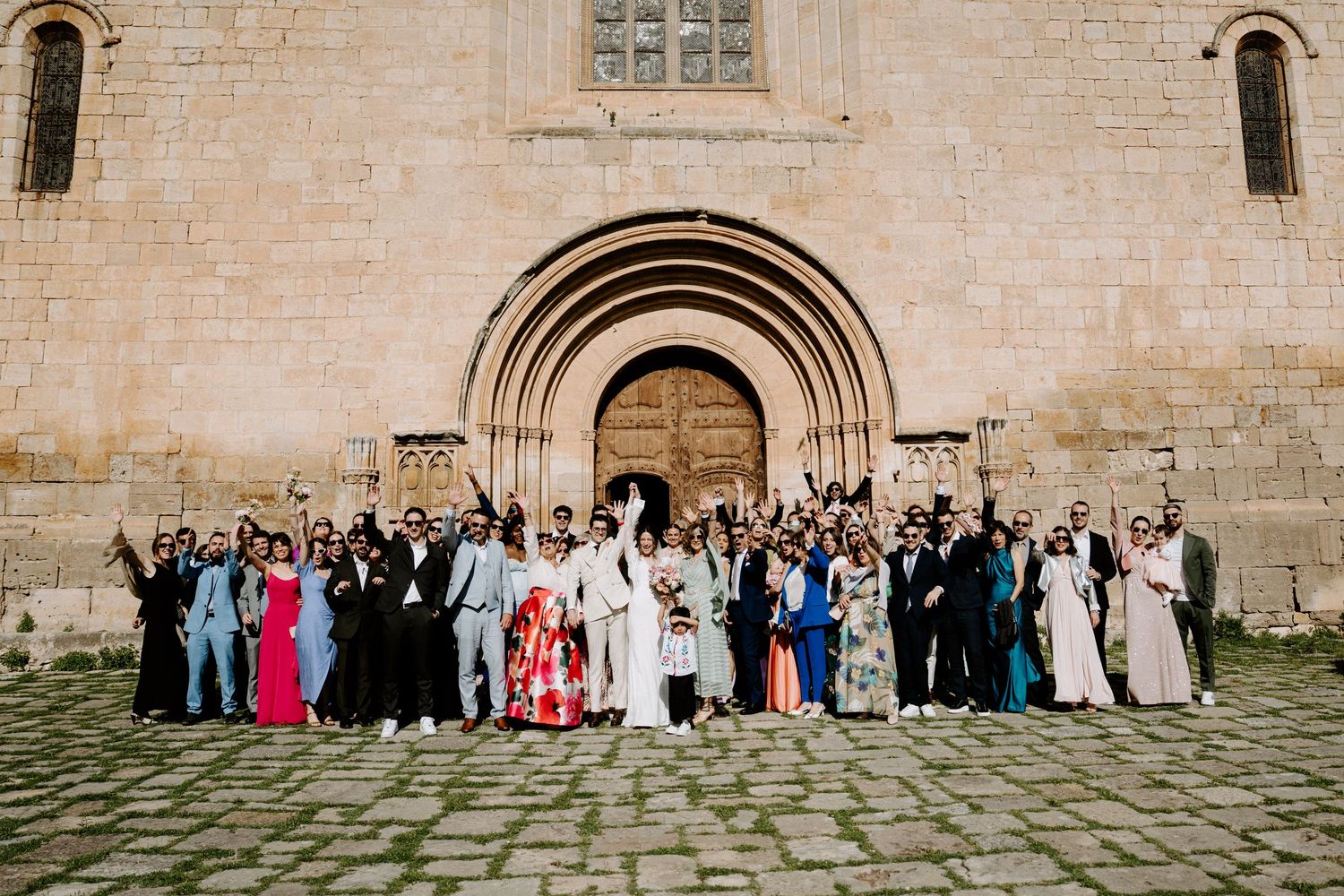 Large wedding party gathered outside historic stone church with arched doorways.