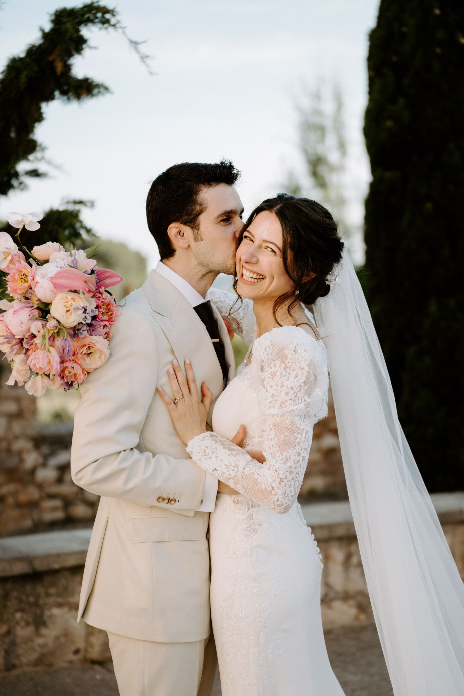 Newlyweds embracing outdoors with pink floral bouquet and flowing veil.