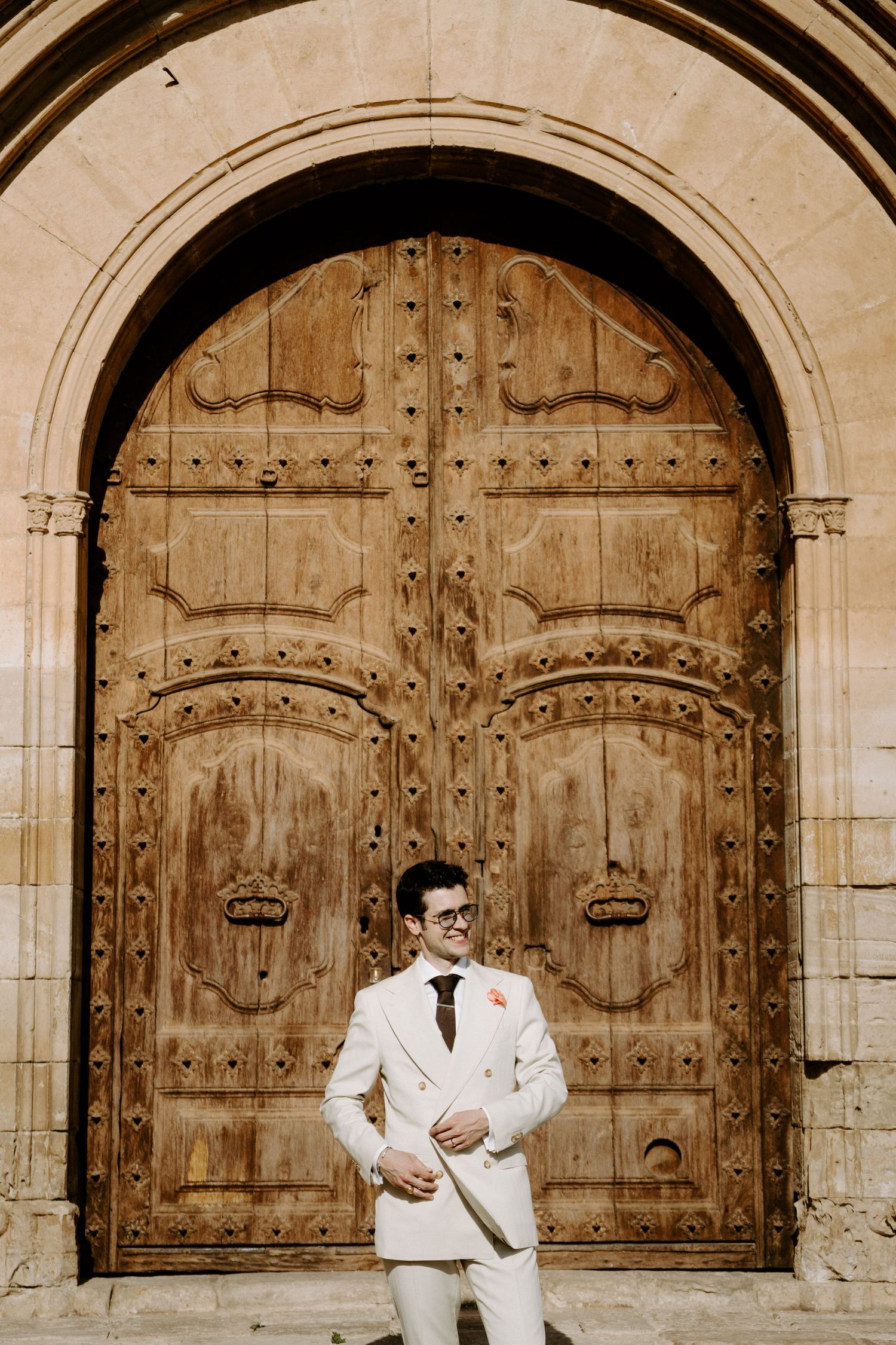 A couple in wedding attire poses in front of ornate wooden cathedral doors.