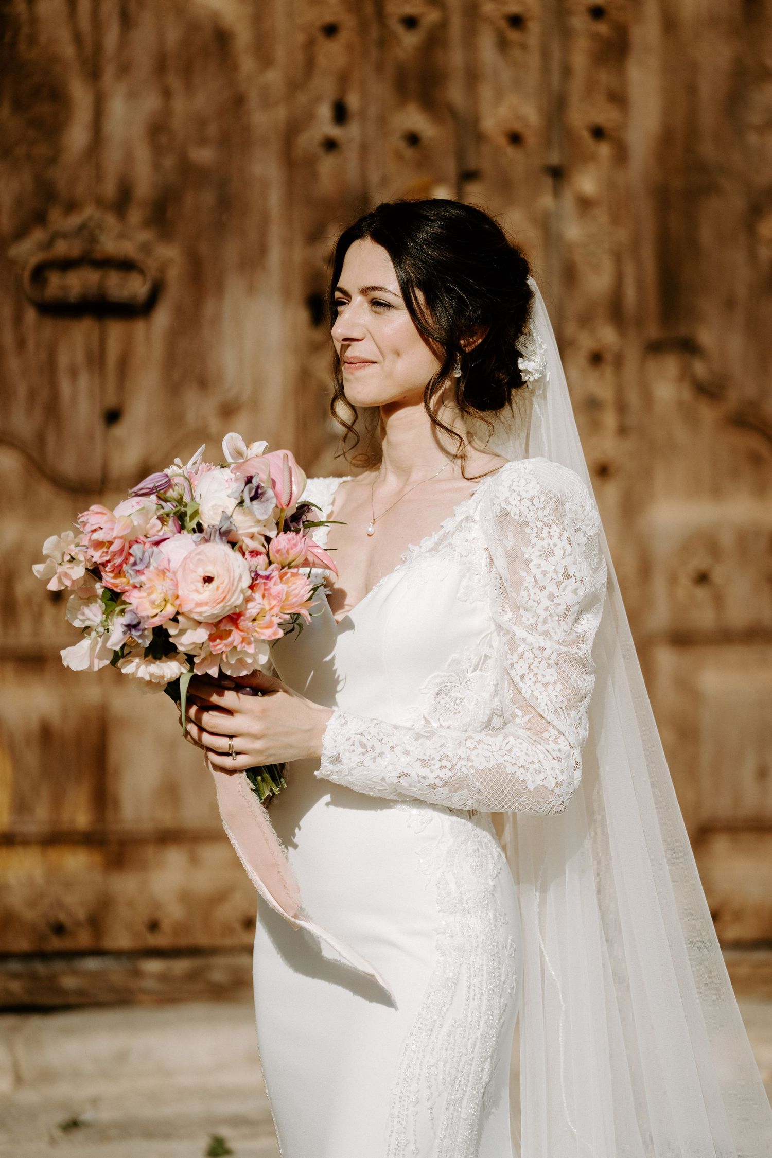 A bride in a lace dress holds a pink and white bouquet against a rustic wooden backdrop.