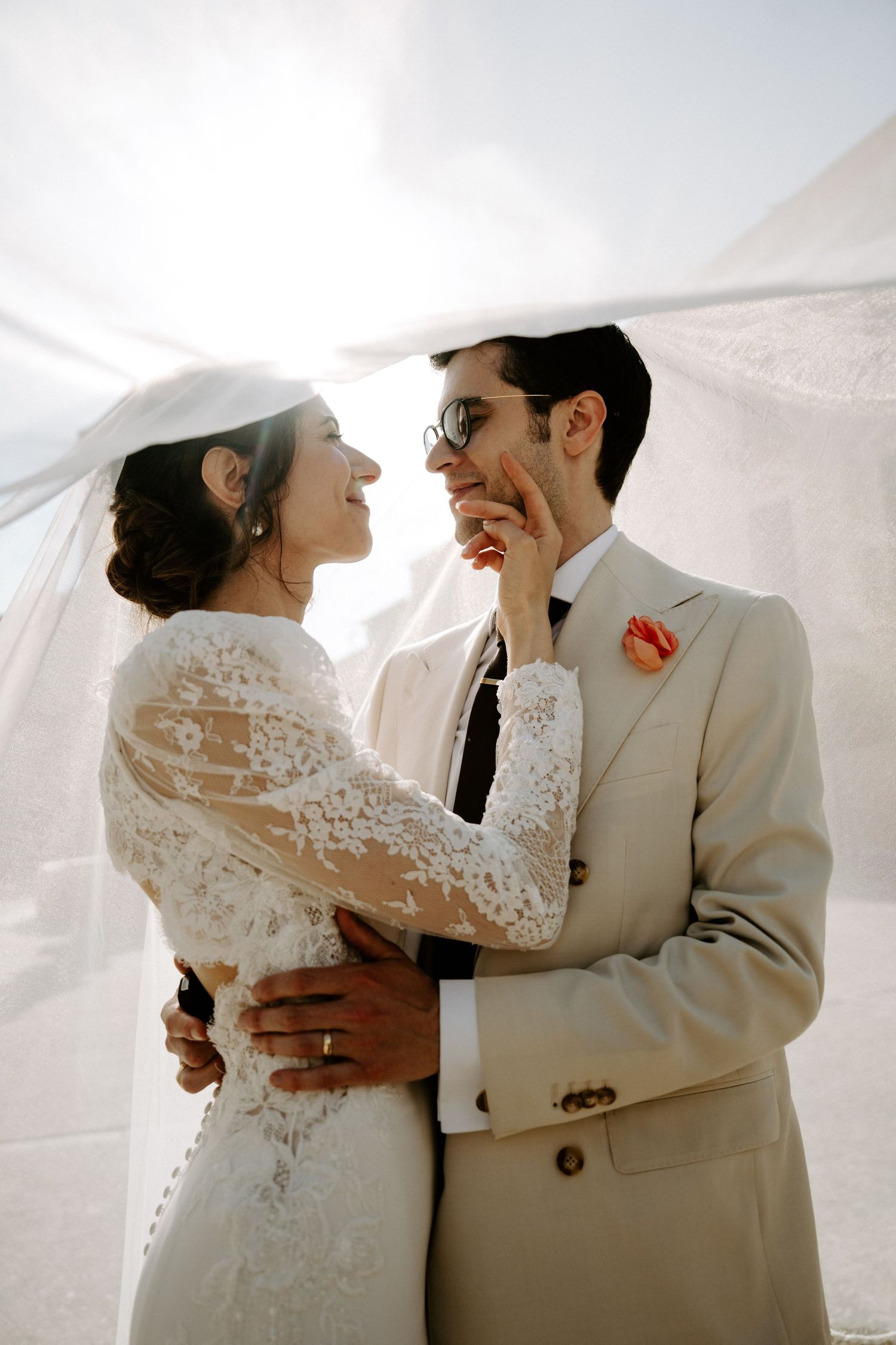 A couple embraces in a sunlit moment wearing elegant white and cream wedding attire.