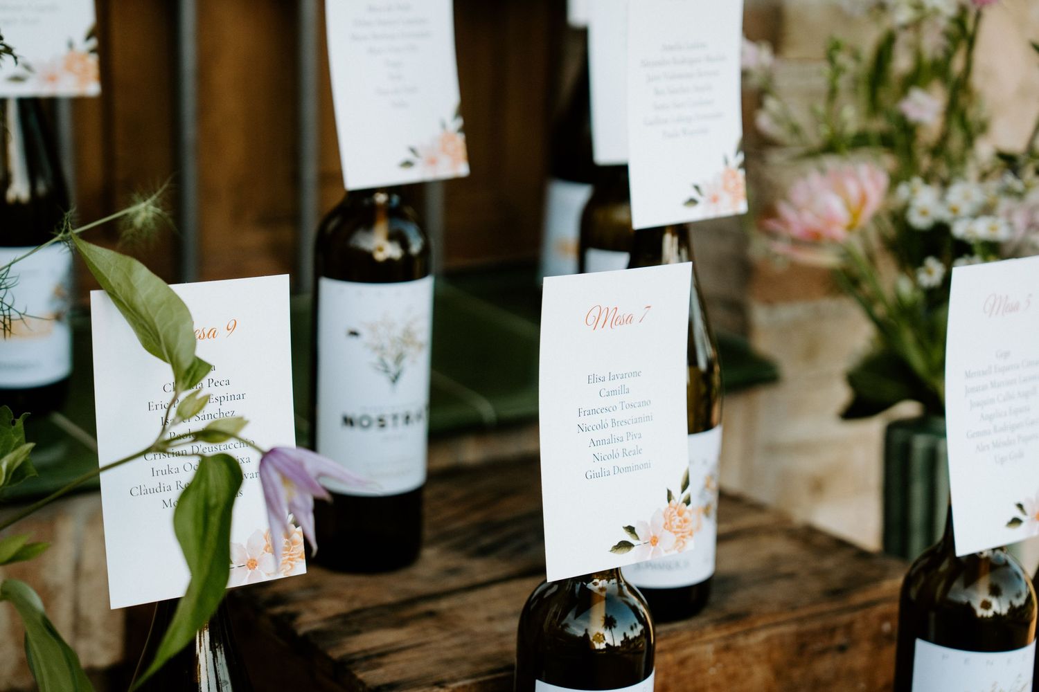Wedding reception table setup with wine bottles and floral arrangements on dark wooden surface.