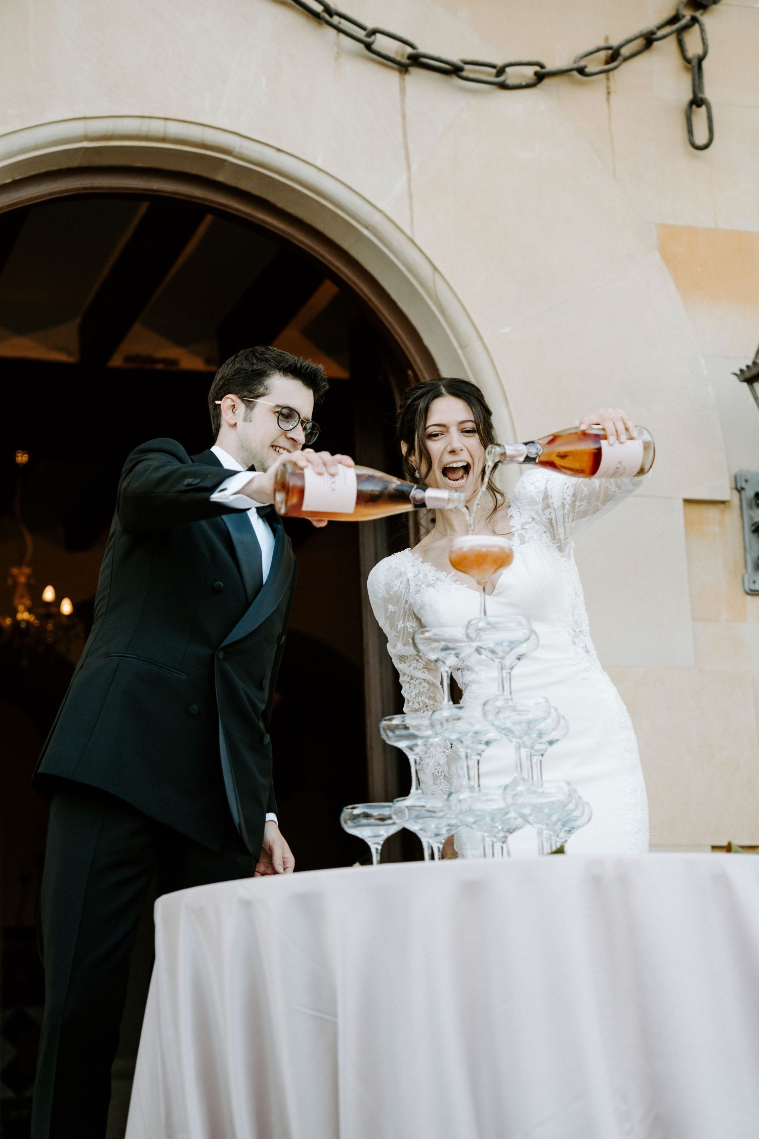 A couple performs a champagne tower pour at an elegant outdoor venue with arched doorways.
