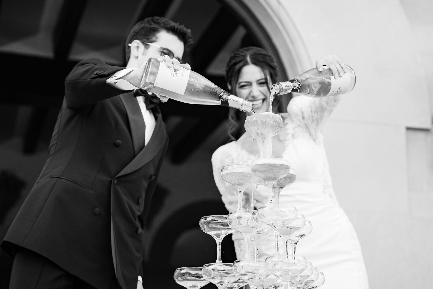 Wedding couple celebrates by pouring champagne into crystal glasses at a stone venue.