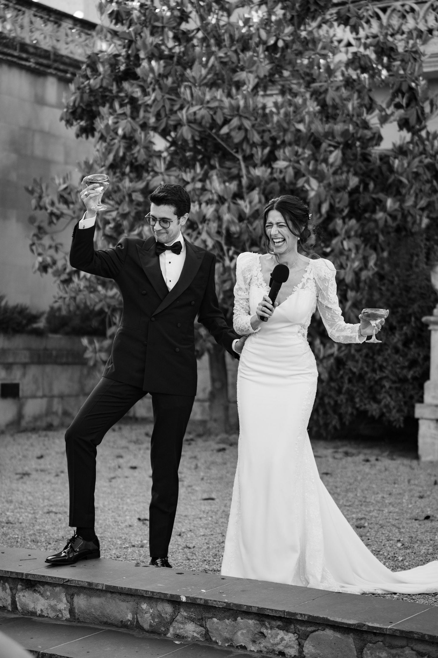 Wedding couple poses playfully in a garden setting with stone walls.