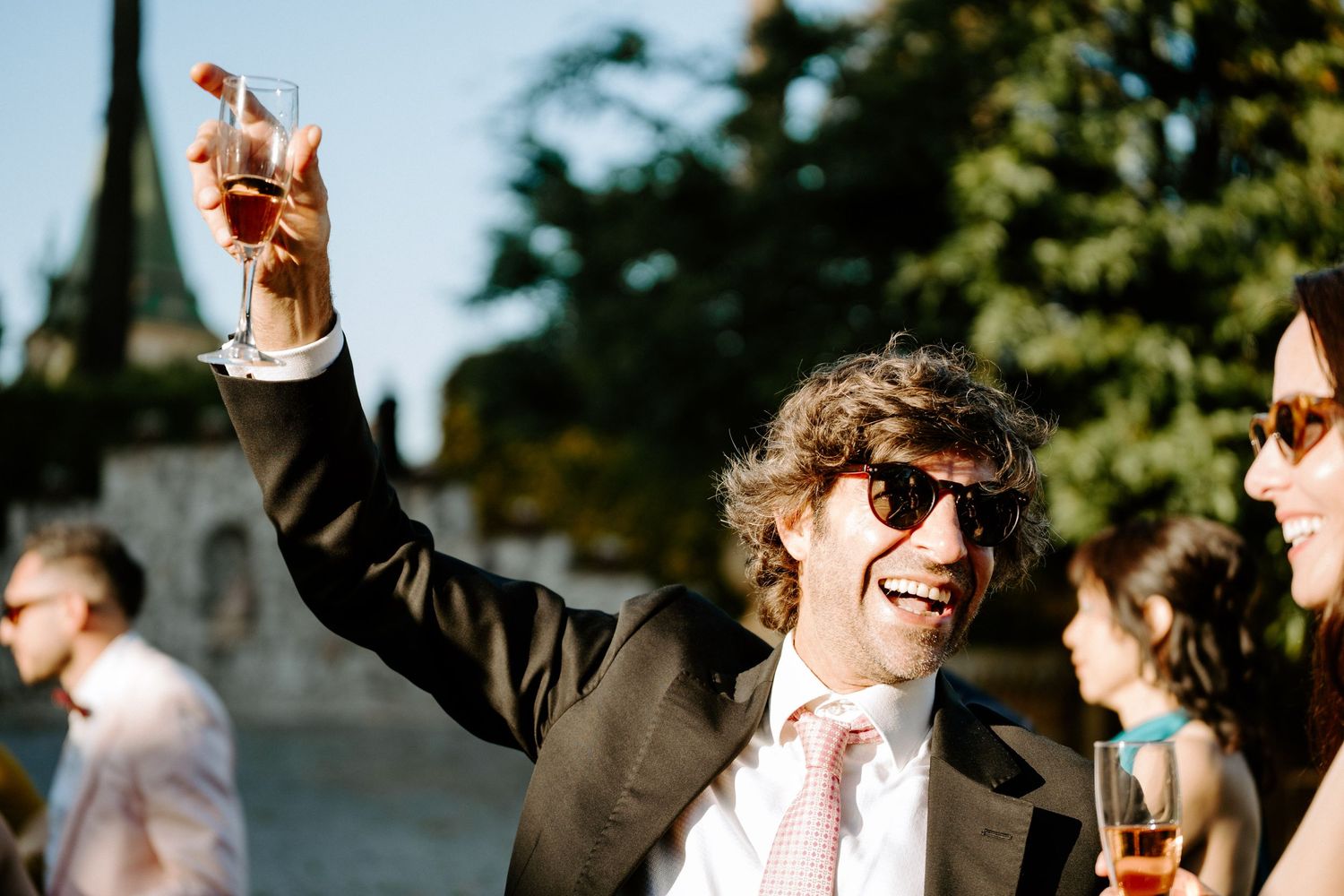 Wedding guests celebrate outdoors with raised glasses in the sunlight.