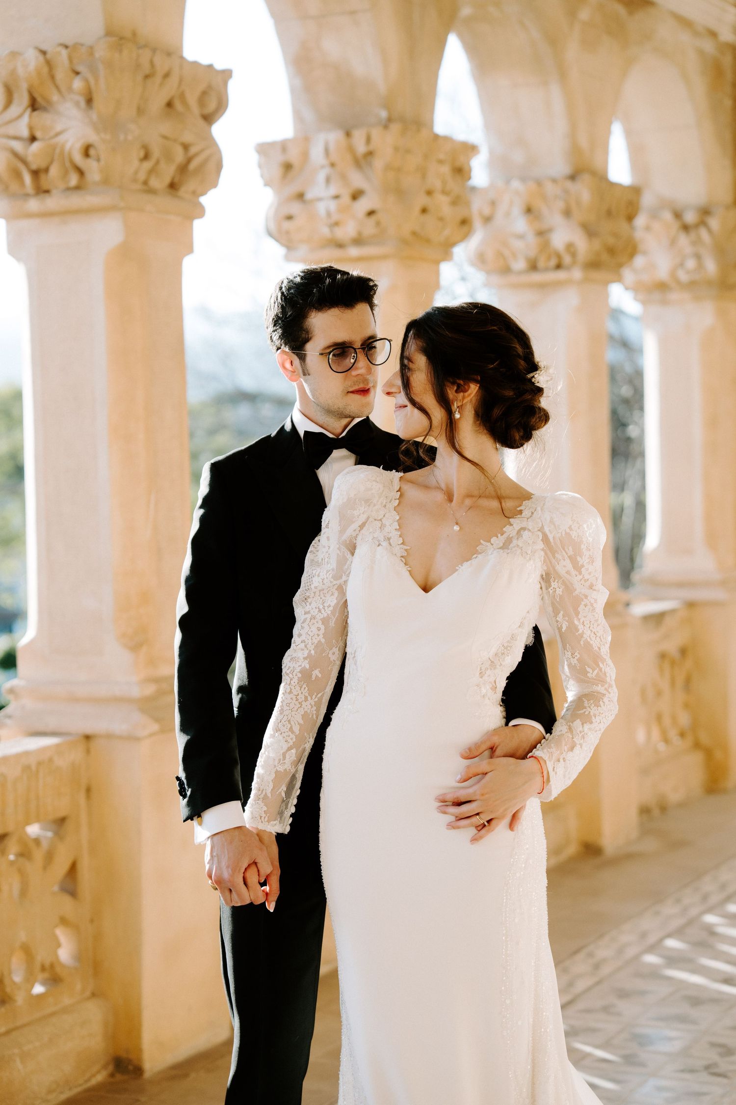 Elegant wedding couple poses in ornate colonnade with decorative stone columns.