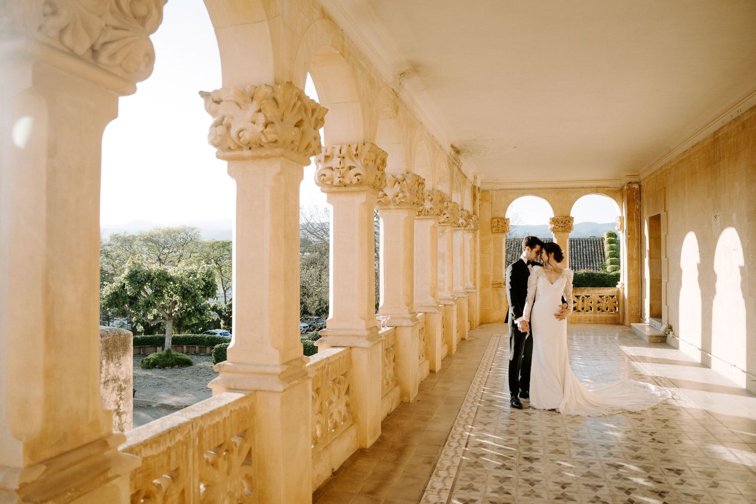 Sunlit colonnade with ornate stone columns creates dramatic wedding venue backdrop.