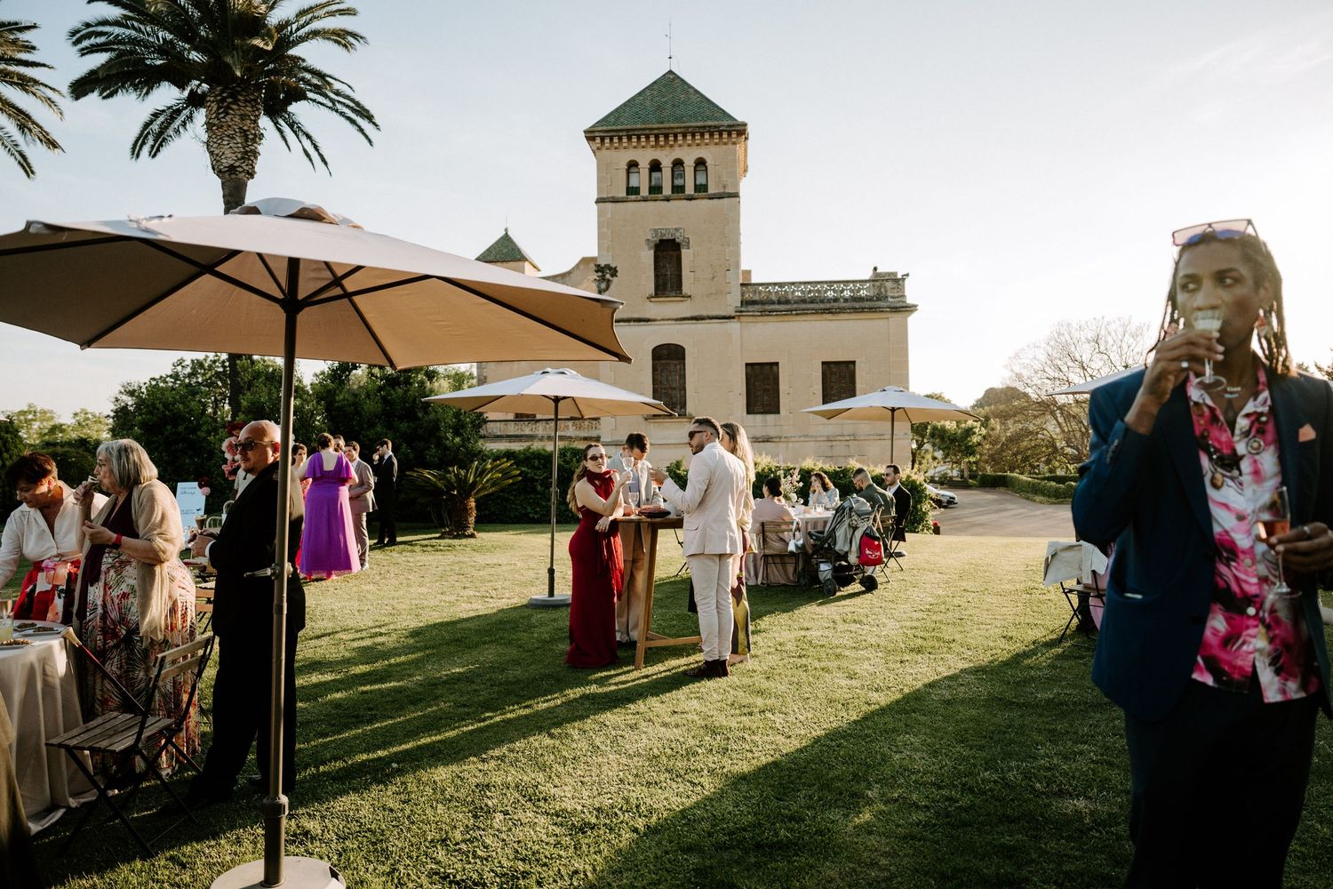 Outdoor wedding reception setup with umbrellas and guests on a grassy lawn.
