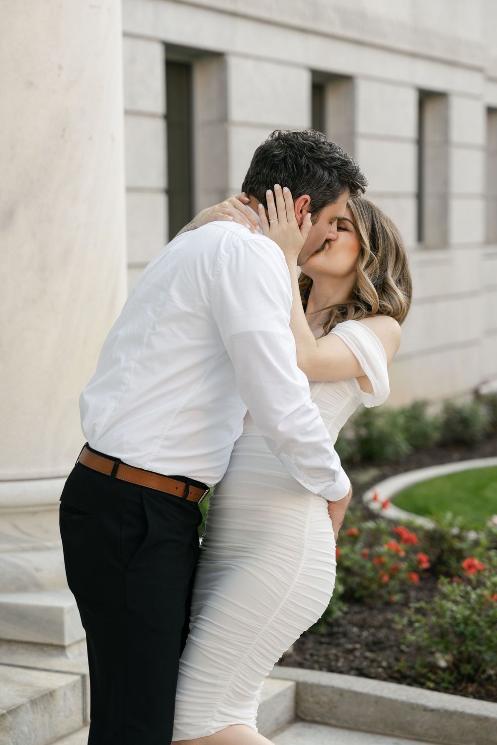 A couple in formal attire sharing a romantic kiss on elegant stone steps outside a building with a garden.