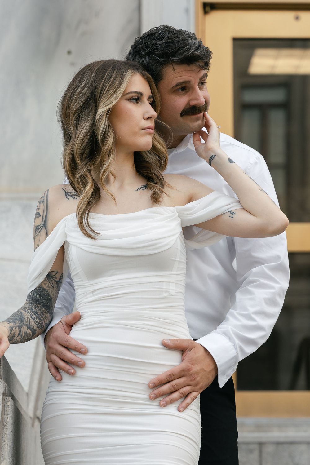 Romantic couple in formal attire poses together against wall, bride in fitted white dress with off-shoulder design.