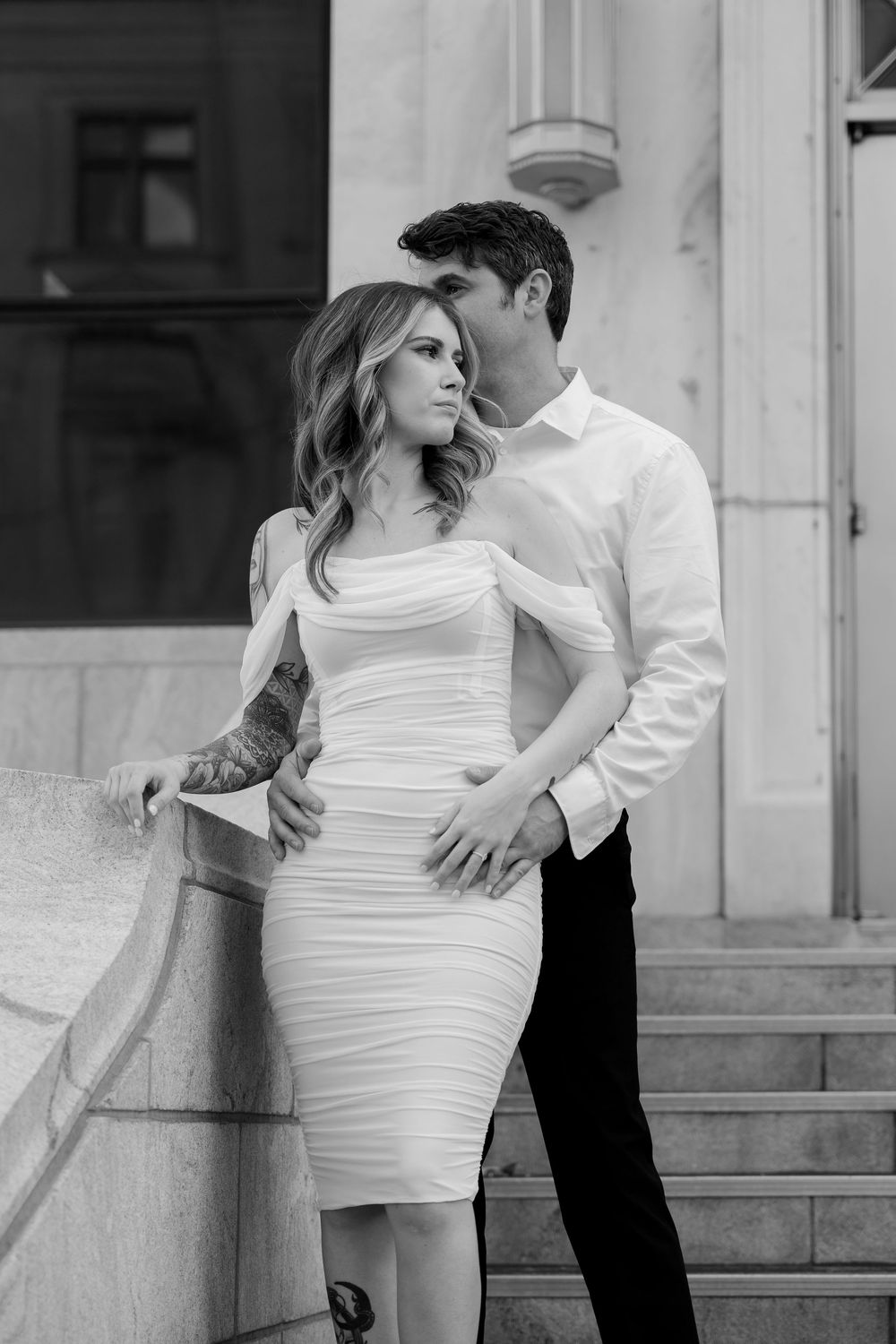Romantic couple in formal attire embraces on stone steps in elegant black and white portrait.