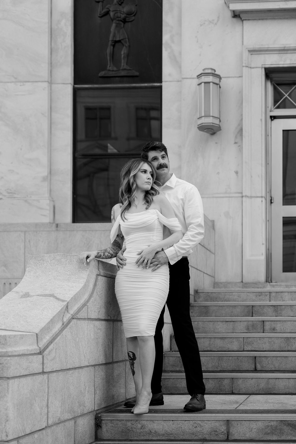 A couple poses romantically on marble steps in front of a white building in black and white photography.