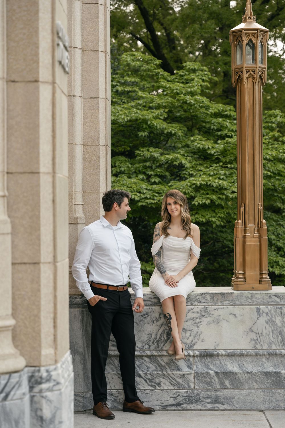 A couple stands together on marble steps near an ornate golden lamp post in an elegant outdoor setting.
