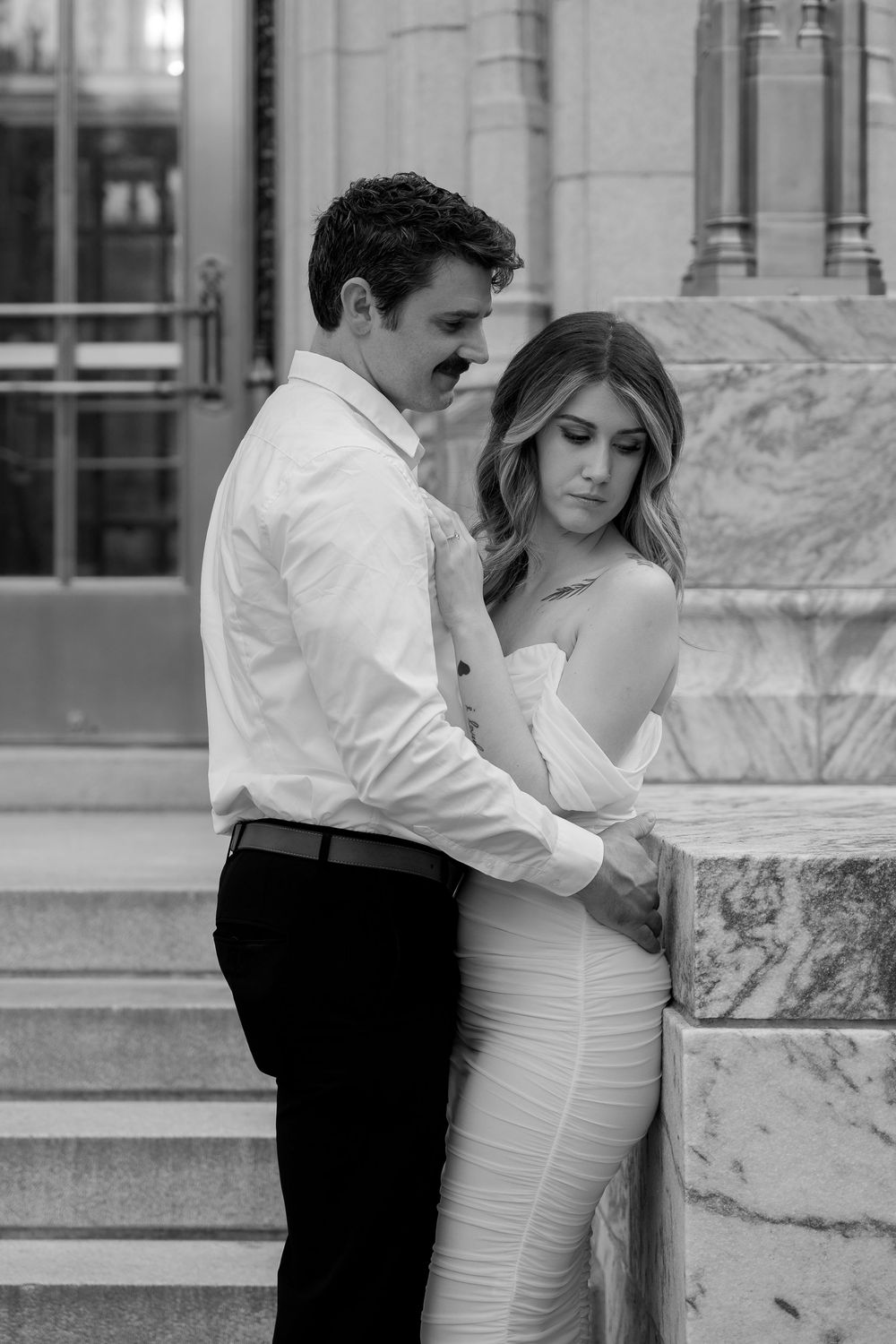 A romantic black and white photo of a couple embracing on marble steps, with the woman in a fitted white dress.