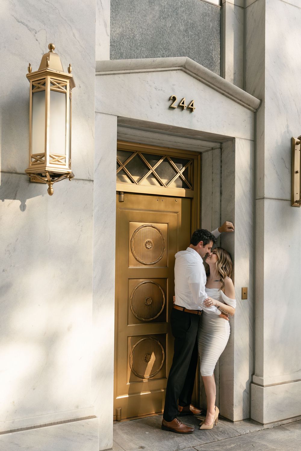 A couple embraces beside an elegant brass door marked 244 with decorative gold sconces on a white building exterior.