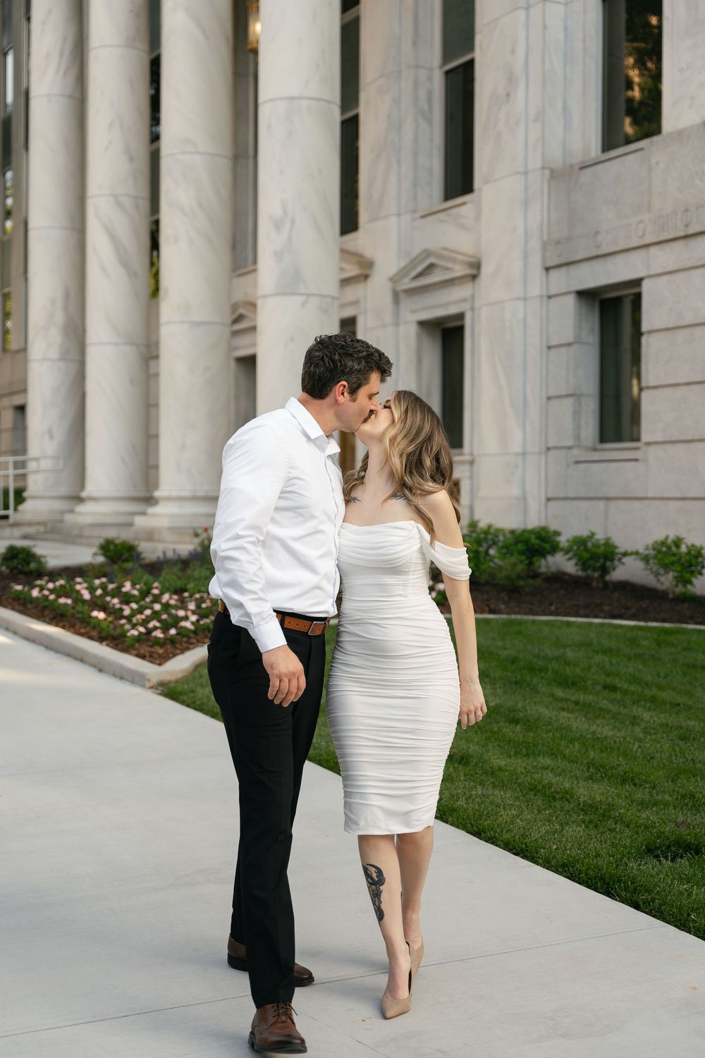 A couple shares a romantic moment outside a stately building with white columns and manicured lawn.
