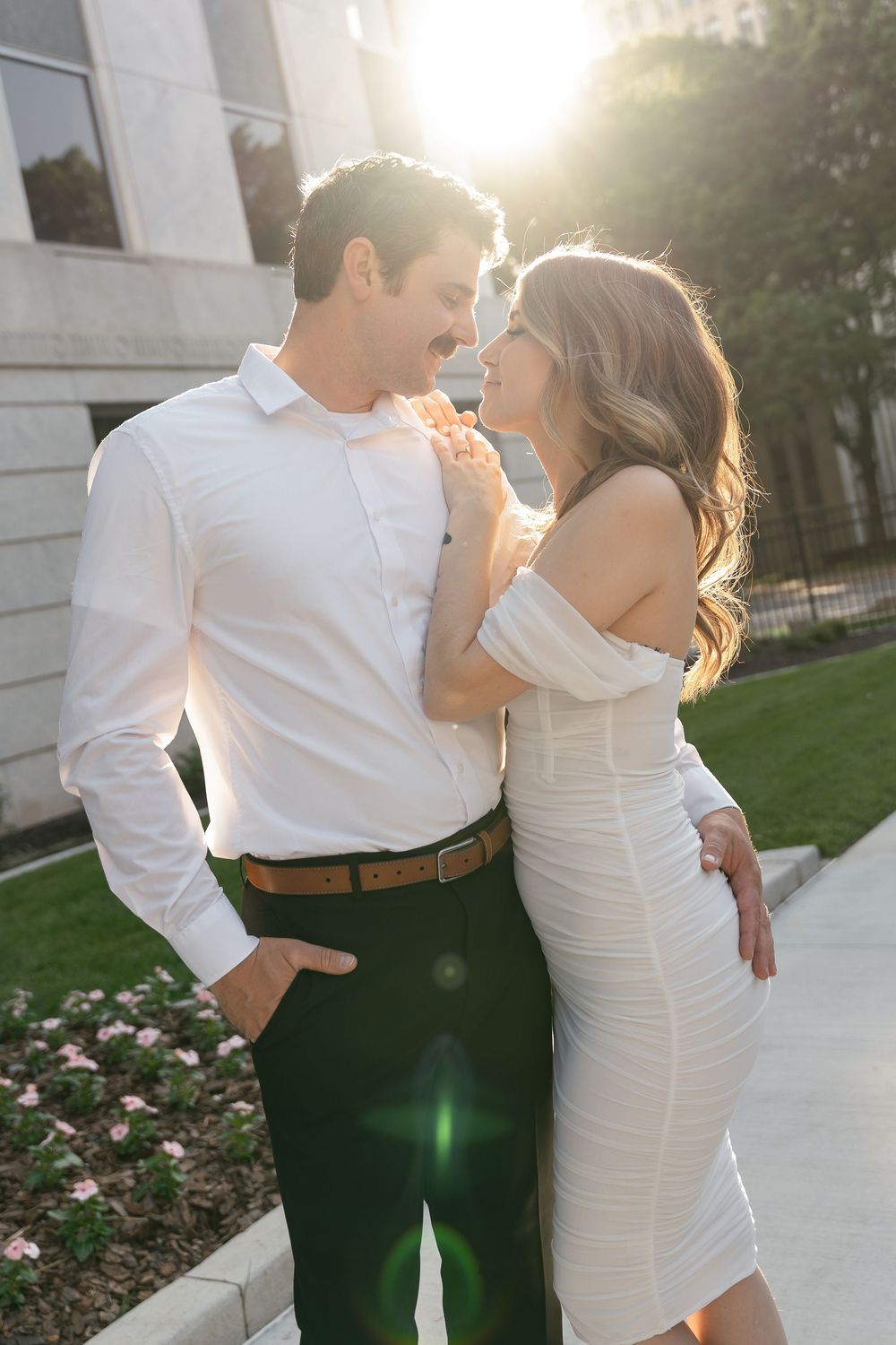 A romantic couple dressed in white embraces outdoors at sunset with a sun flare in the background.