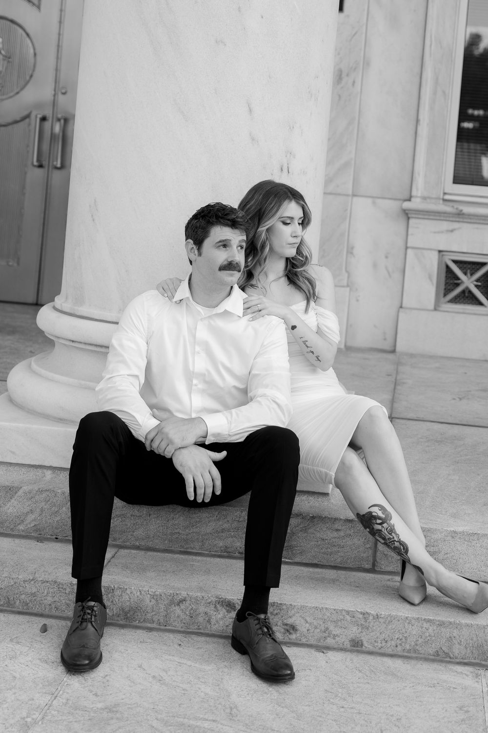 A romantic black and white photo of a couple sitting on stone steps in elegant white and black attire.