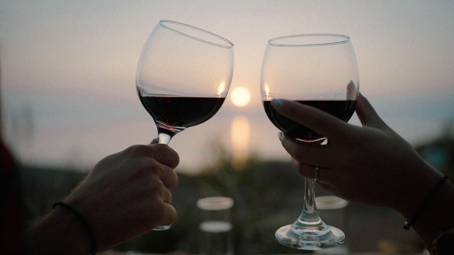 Two wine glasses clinking together at sunset with blurred background lighting creating a romantic silhouette.