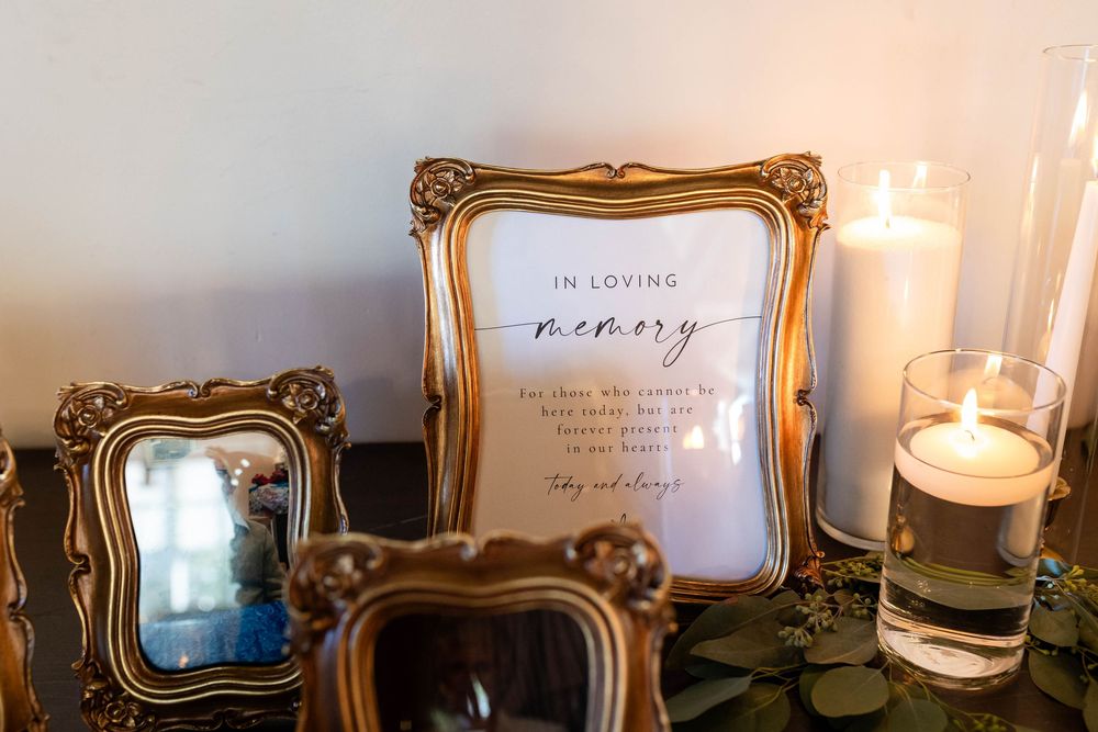 Vintage-style gold frames and memorial candle display with 'In Loving Memory' text on a dark table surface.