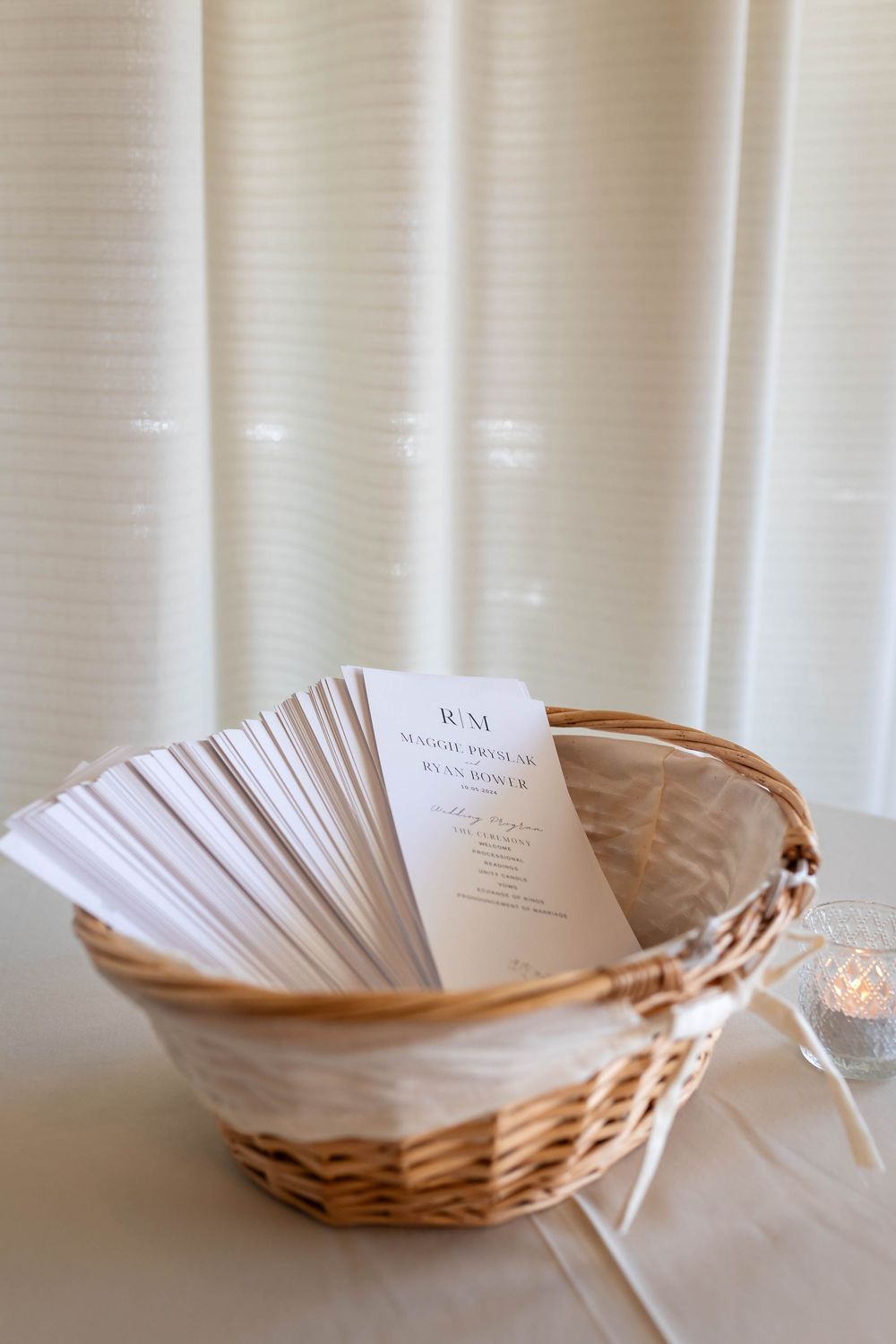 A woven basket filled with white paper wedding programs sits against a light curtain background.