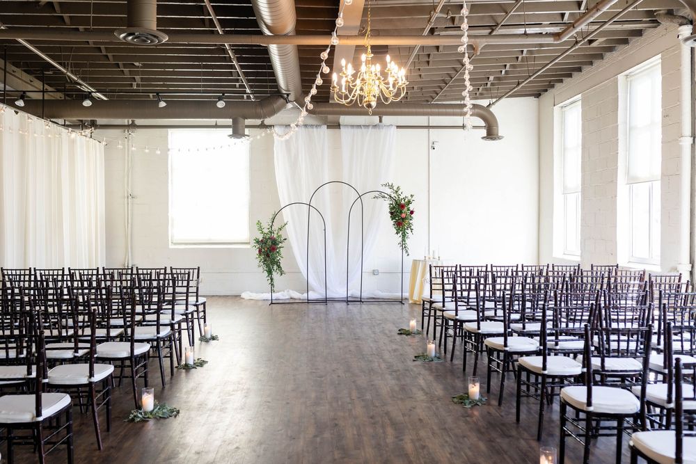 Modern wedding ceremony venue with white chairs, arched altar, and elegant chandelier in an industrial-style loft space.