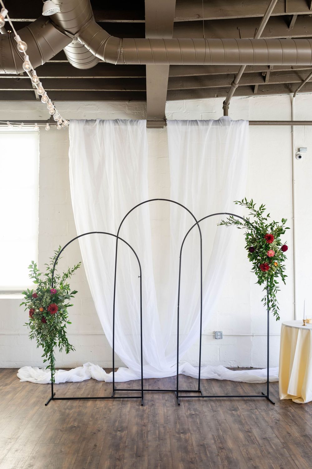 Modern wedding arch setup with black metal frames, white draping, and red floral arrangements on hardwood floor.