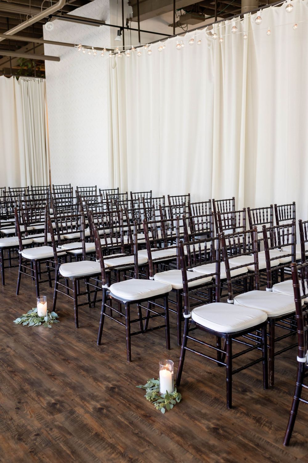 Rows of dark Chiavari chairs with white cushions arranged for a wedding ceremony with white draping and candle decor.