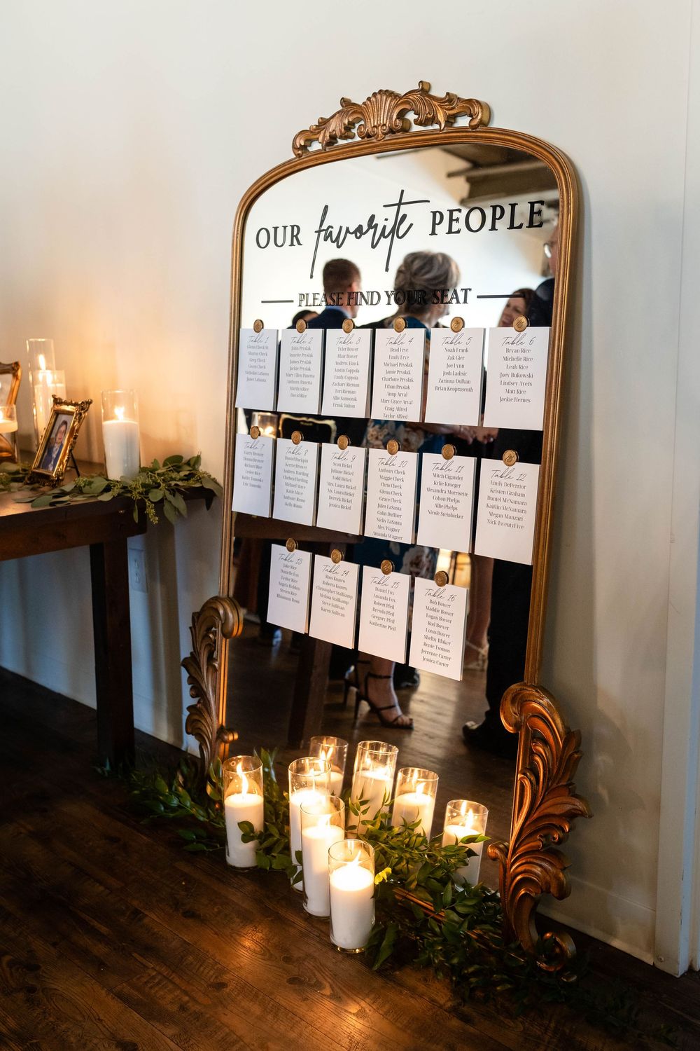 An ornate gold mirror displays a wedding seating chart with lit candles and greenery at the base.
