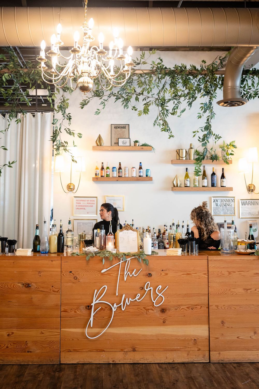 A rustic wooden bar with neon signage, crystal chandelier, and floating shelves displaying bottles under greenery decor.