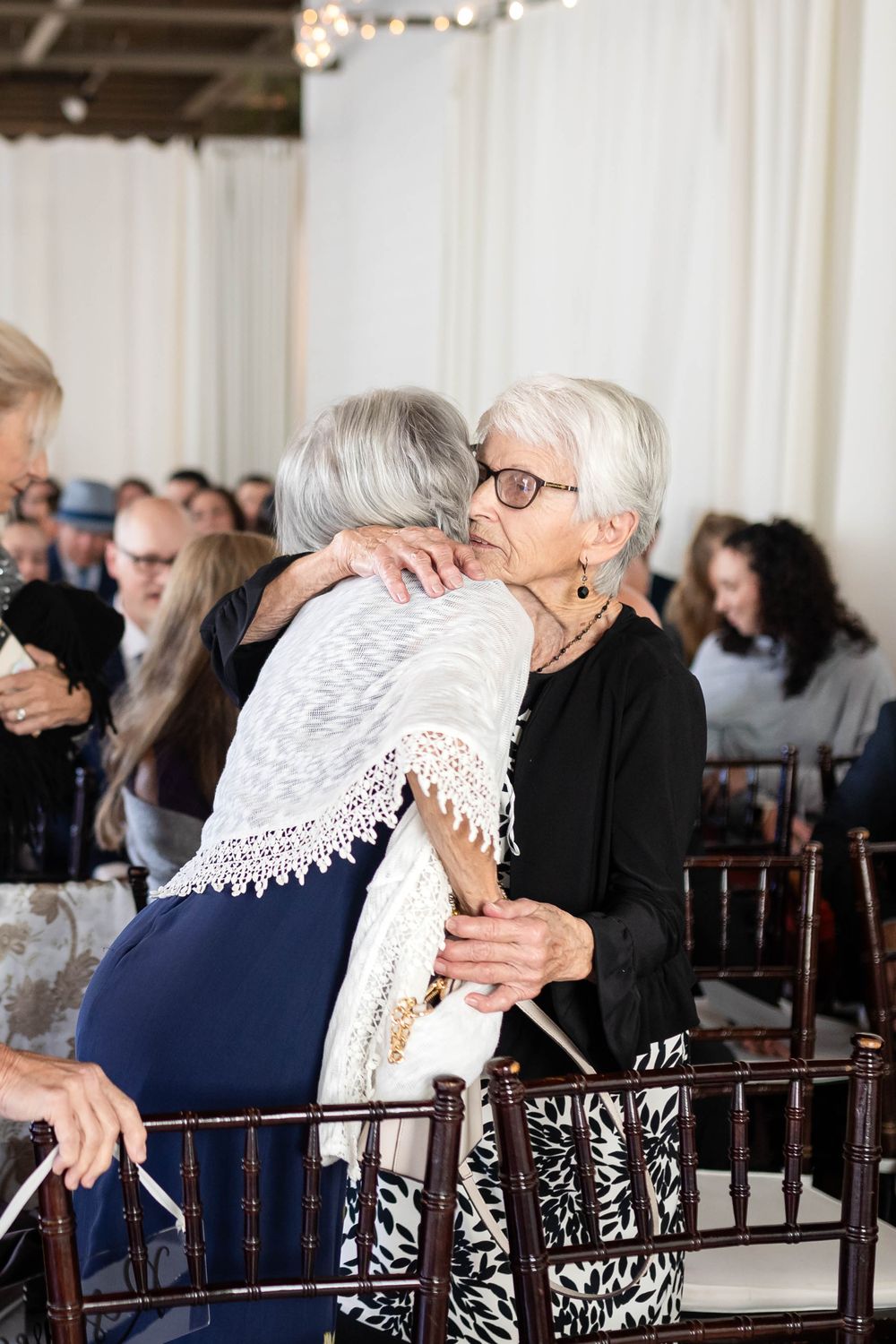 Two elderly women share a warm embrace at an indoor event with banquet chairs and white drapery in the background.