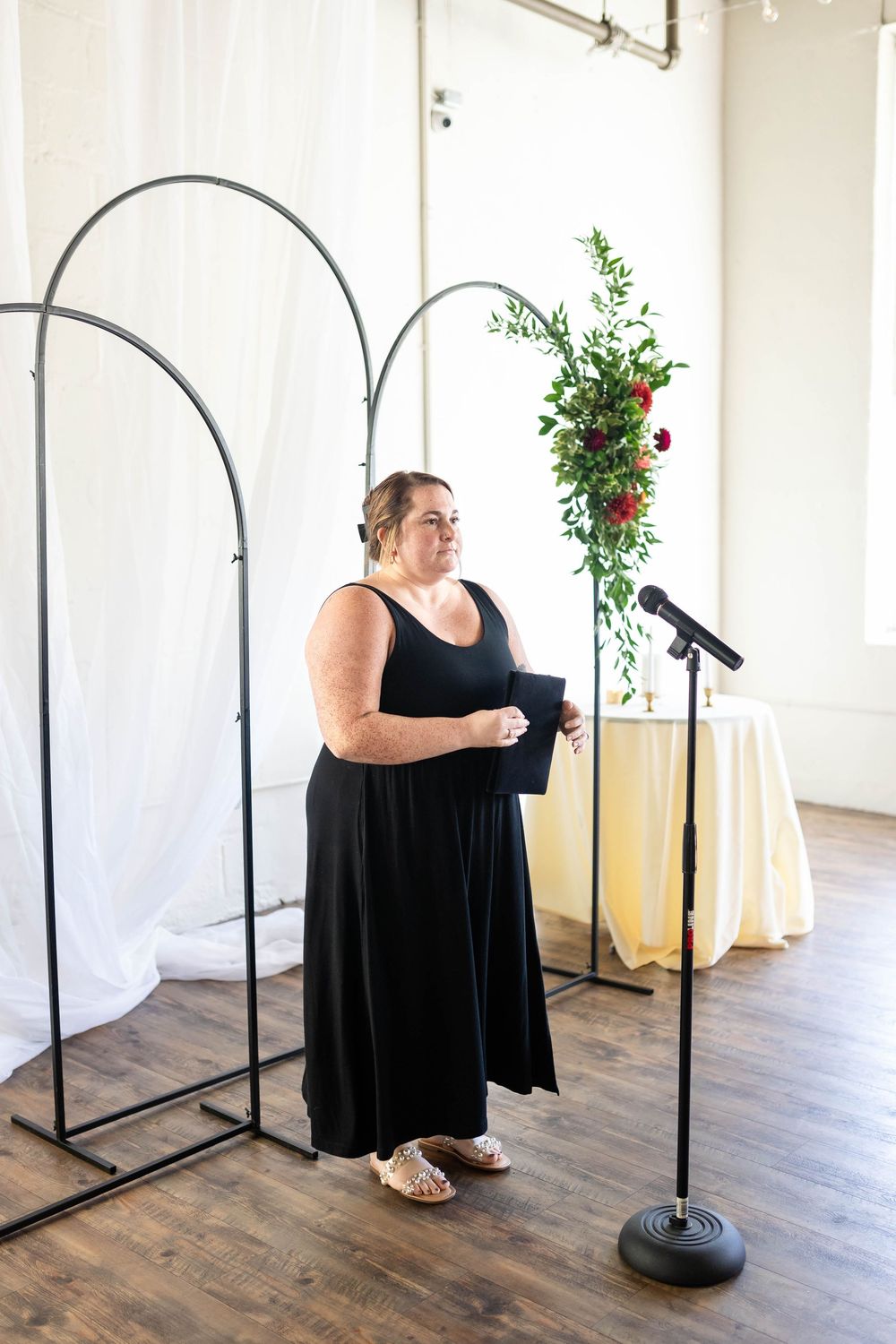 A person in a black dress stands at a microphone in a light-filled room with arched metal frames and floral decor.