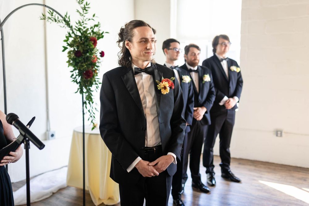 Groomsmen in black tuxedos stand in line at an indoor wedding venue with floral decorations.