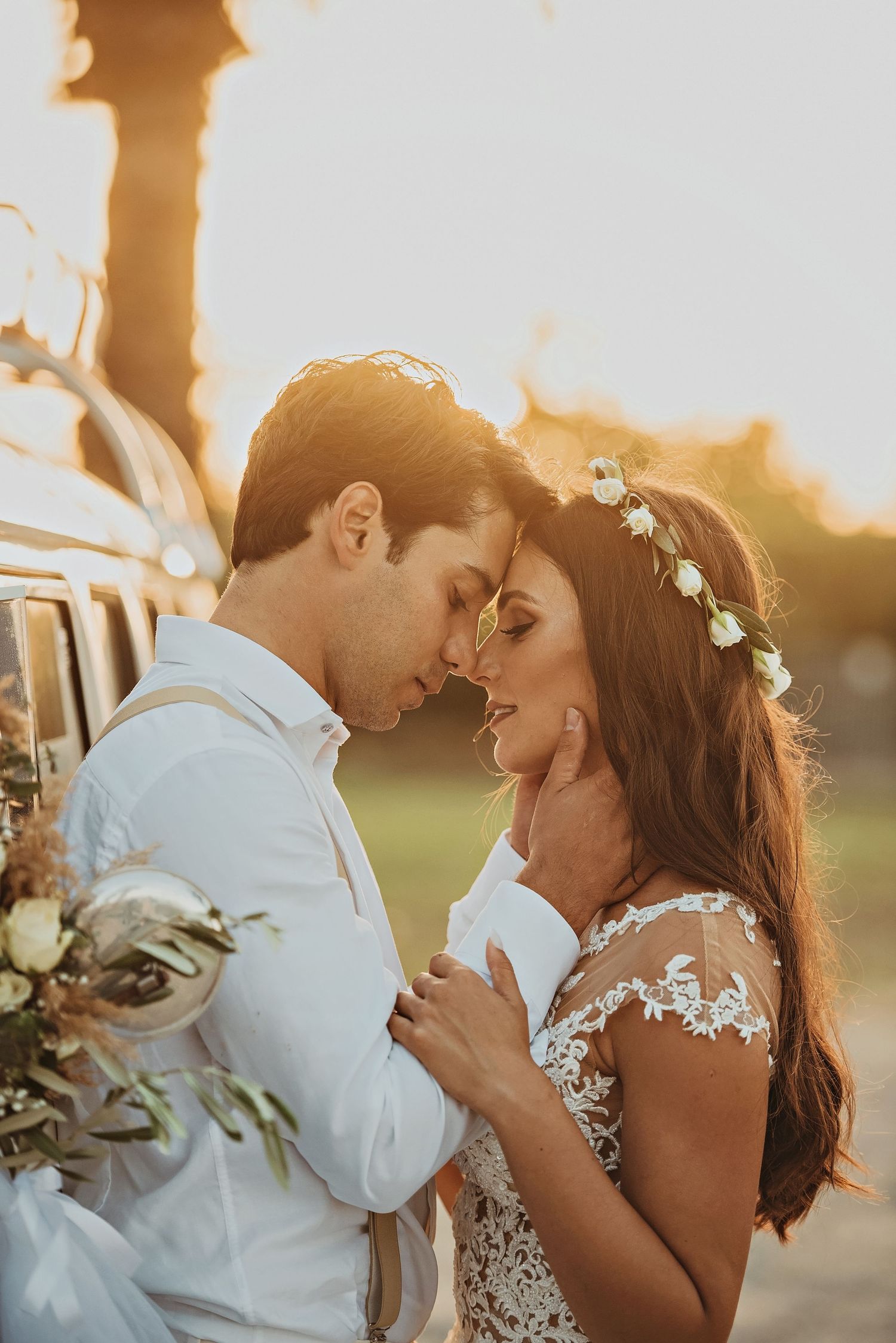 A bride and groom ride skateboards together on a tree-lined road during sunset in their wedding attire.