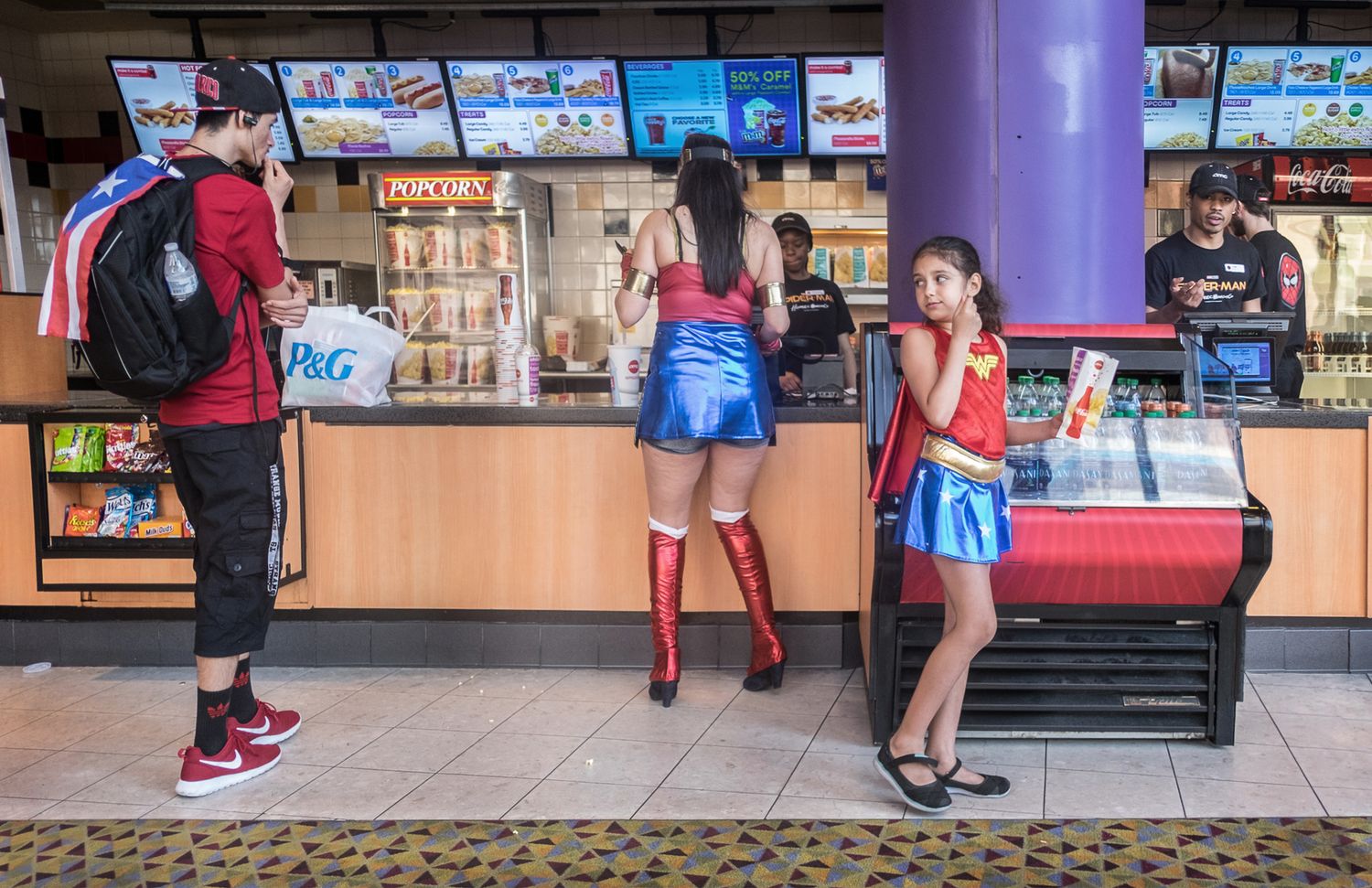 People in colorful costumes stand at a concession counter with multiple menu screens displayed above.
