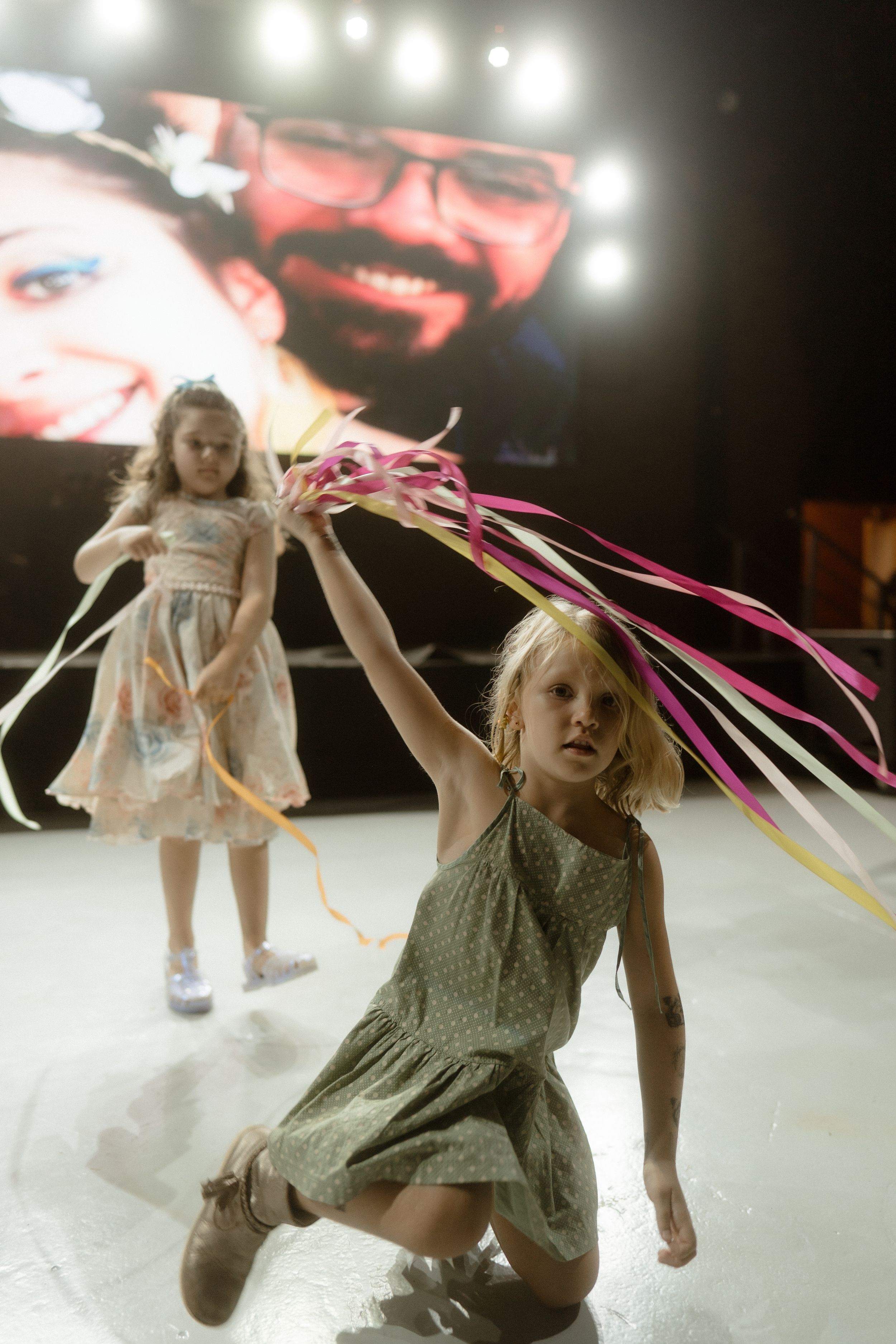 Young dancers perform in white dresses with colorful ribbons on a stage under bright spotlights.