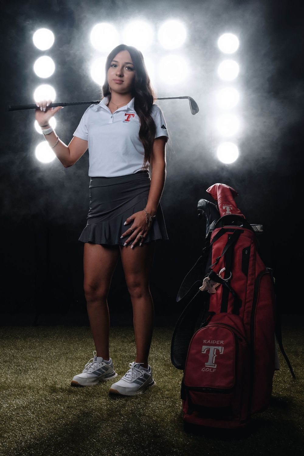Dramatic portrait of a golfer in athletic wear standing with club next to red golf bag with spotlights in background.