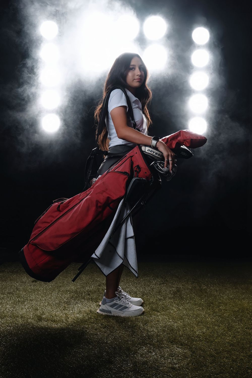 Athletic golfer with red golf bag stands on grass field at night with dramatic spotlights and fog in background.