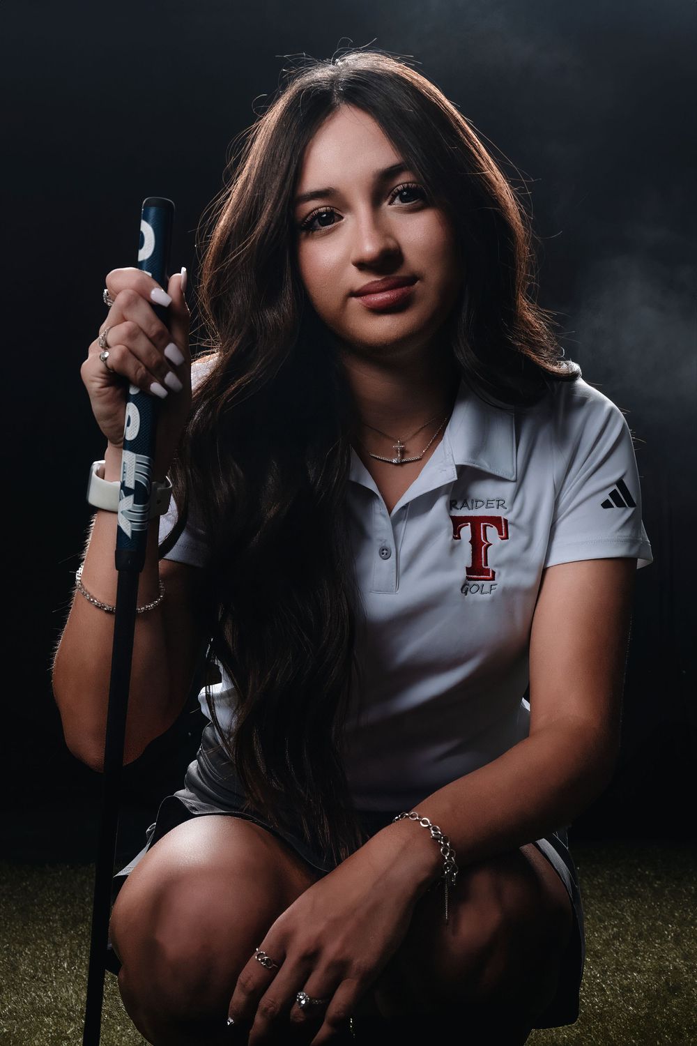 Portrait of an athlete in a white polo with a T logo holding a golf club against a dark background.