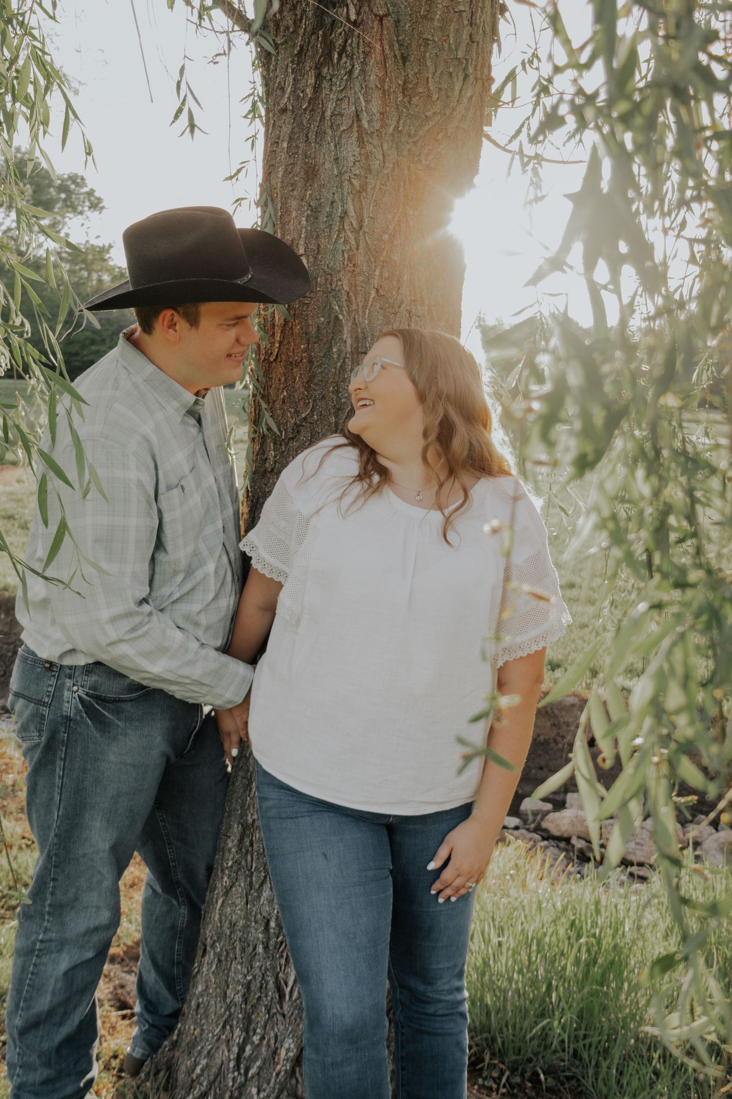 A couple shares a romantic moment leaning against a willow tree in casual western attire during a sunlit afternoon.