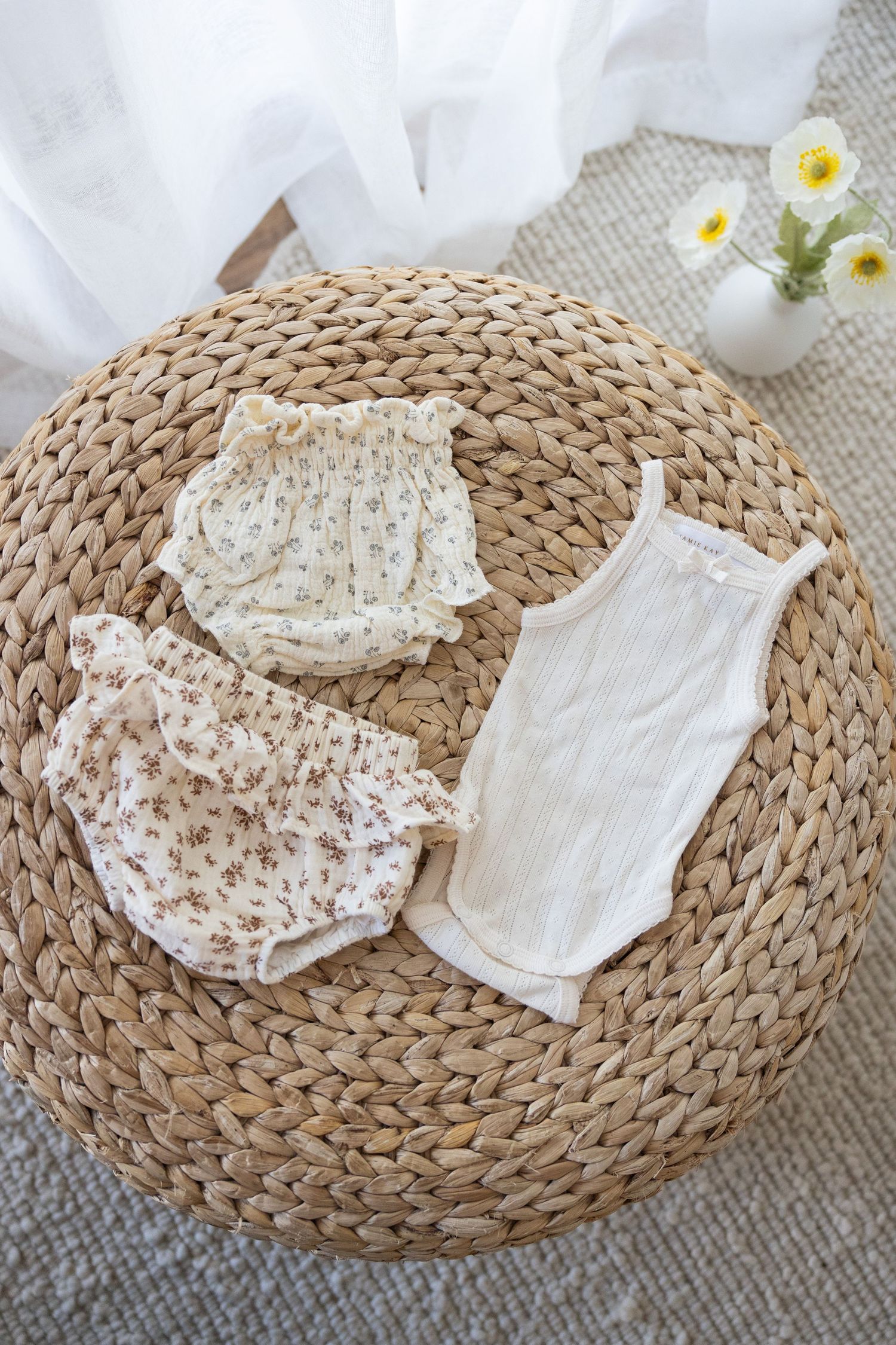 Delicate white baby clothes and undergarments displayed on a woven round rattan placemat with small daisies in vase.