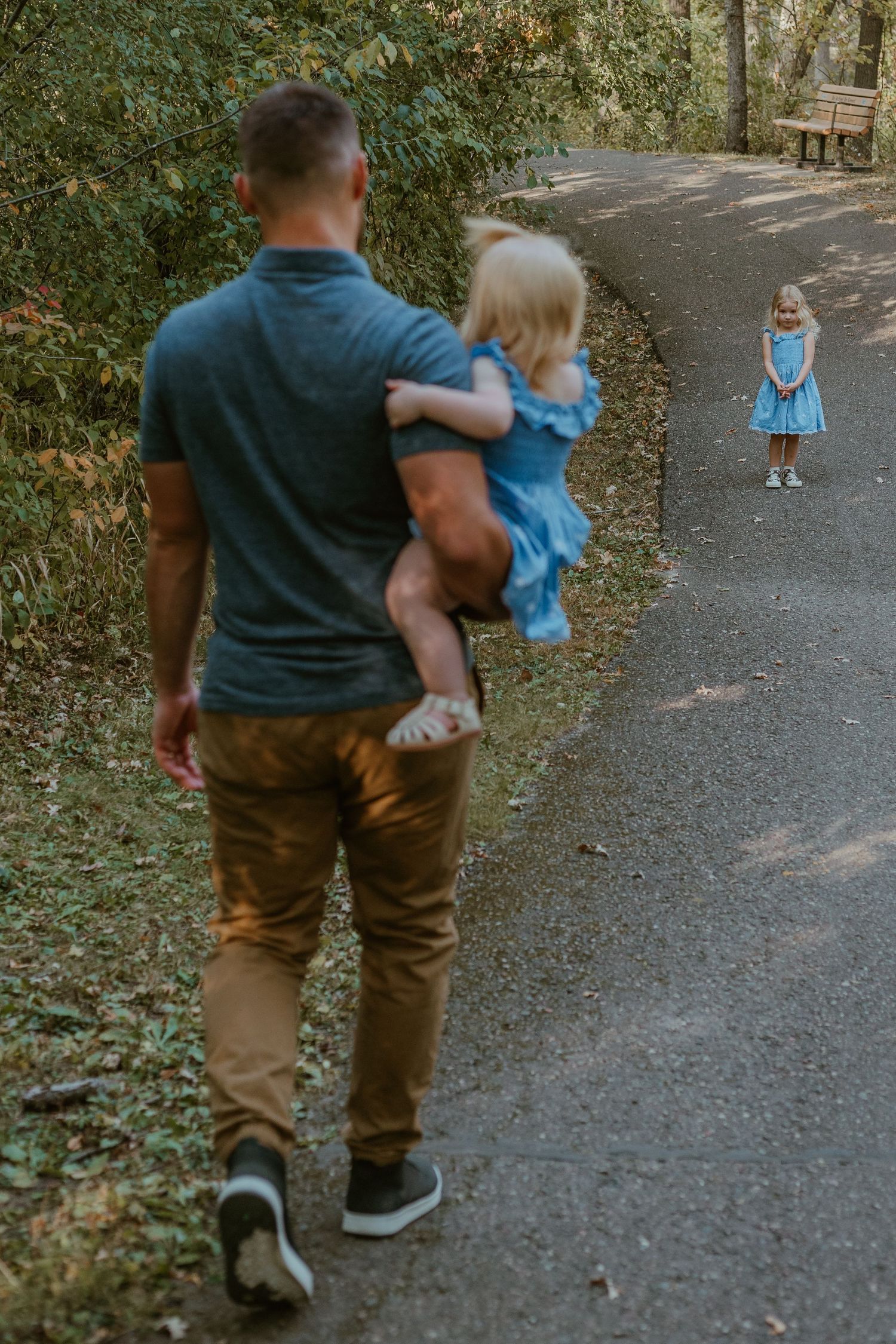 Two children in matching blue dresses walking along a wooded path while being carried by someone in a green shirt.