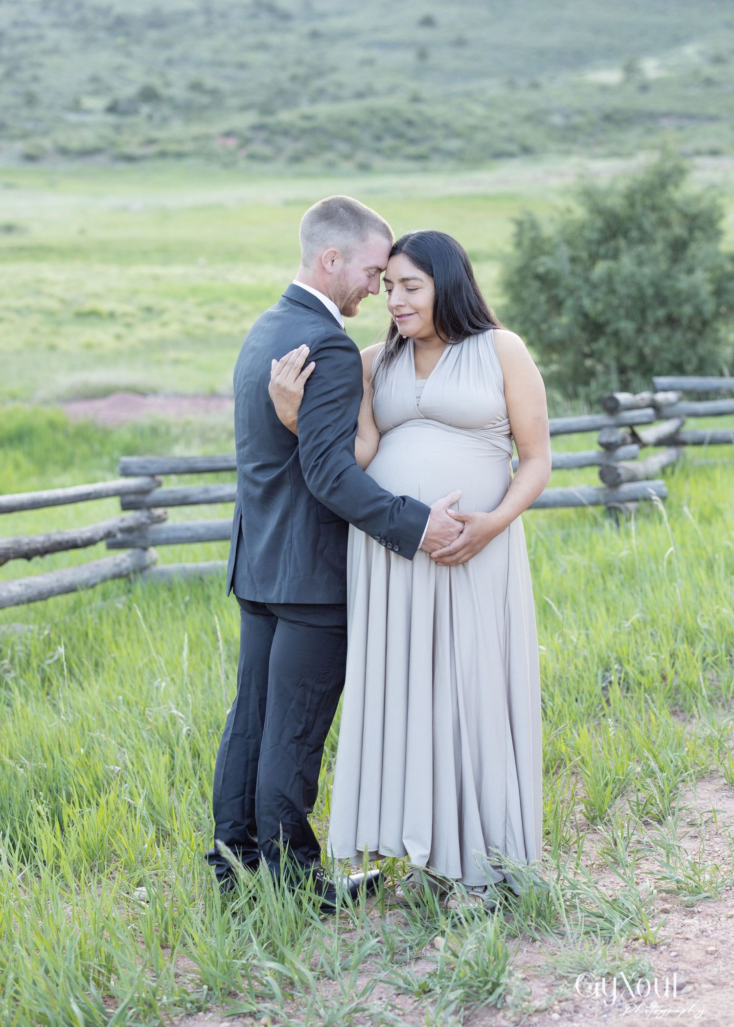 A couple embraces in a scenic field with a rustic wooden fence, green grass and rolling hills in the background.