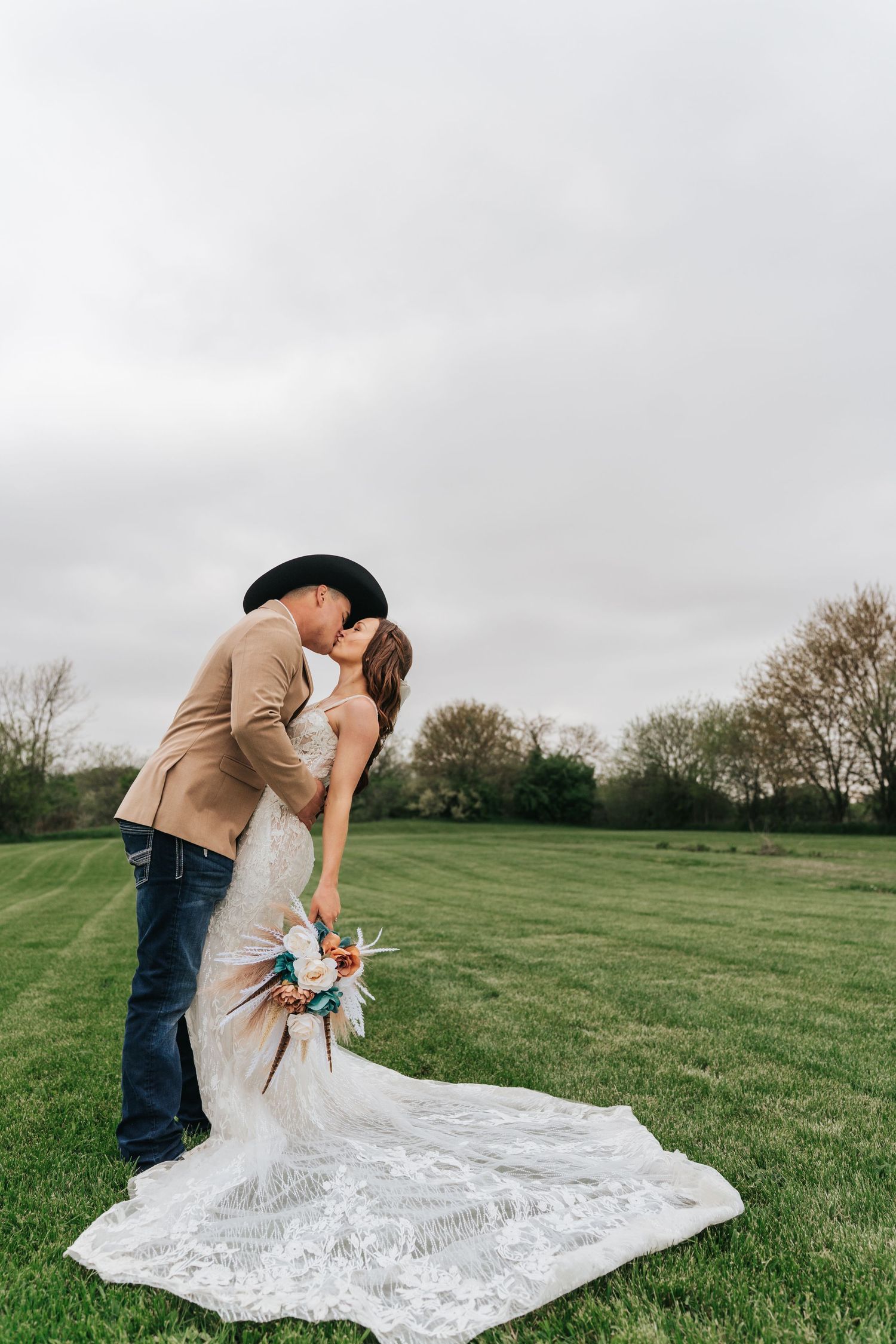 Romantic wedding couple embraces in an open field with long lace train trailing behind.
