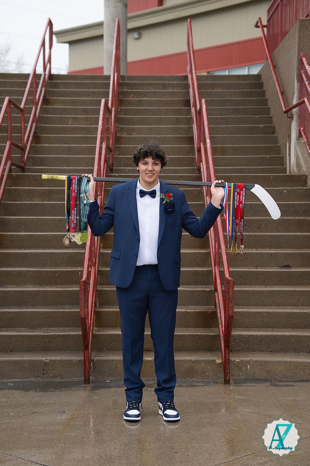 Someone in a navy blue suit and bow tie holds up two vintage wooden skis while standing on concrete stairs with red railings.